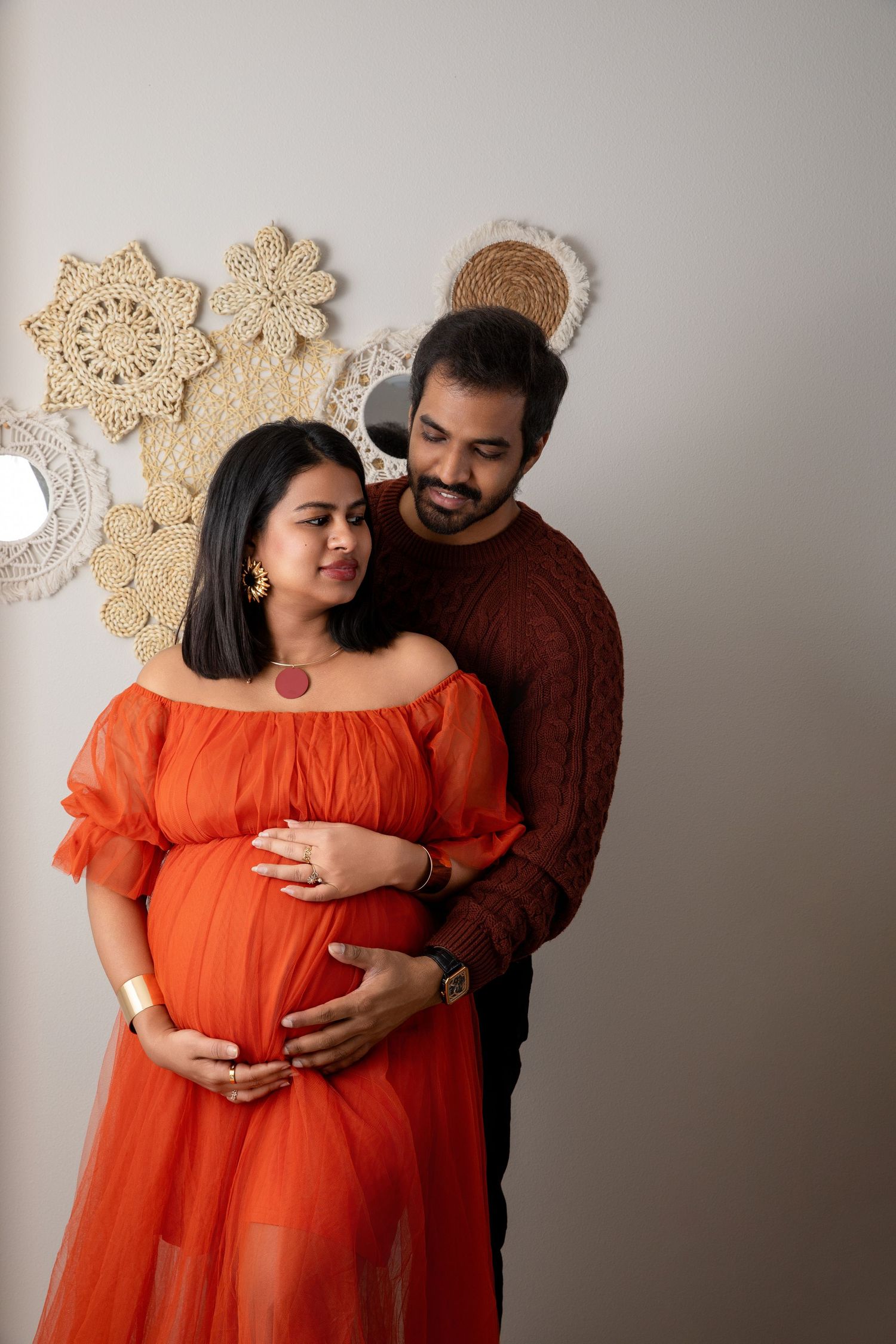 Couple embraces during maternity photoshoot with woman in flowing orange dress.