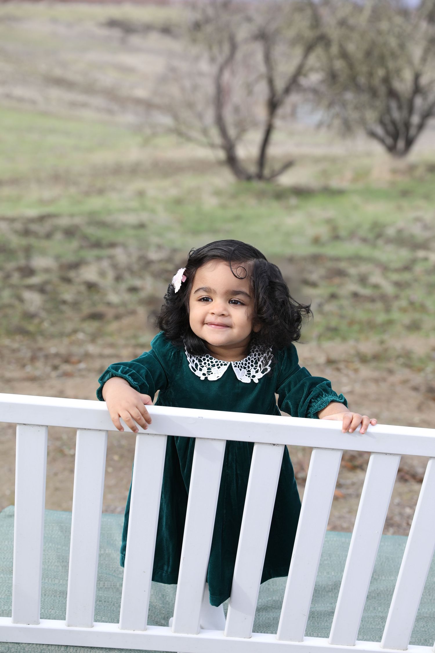 A child in a green velvet dress with white collar leans on a white railing outdoors.