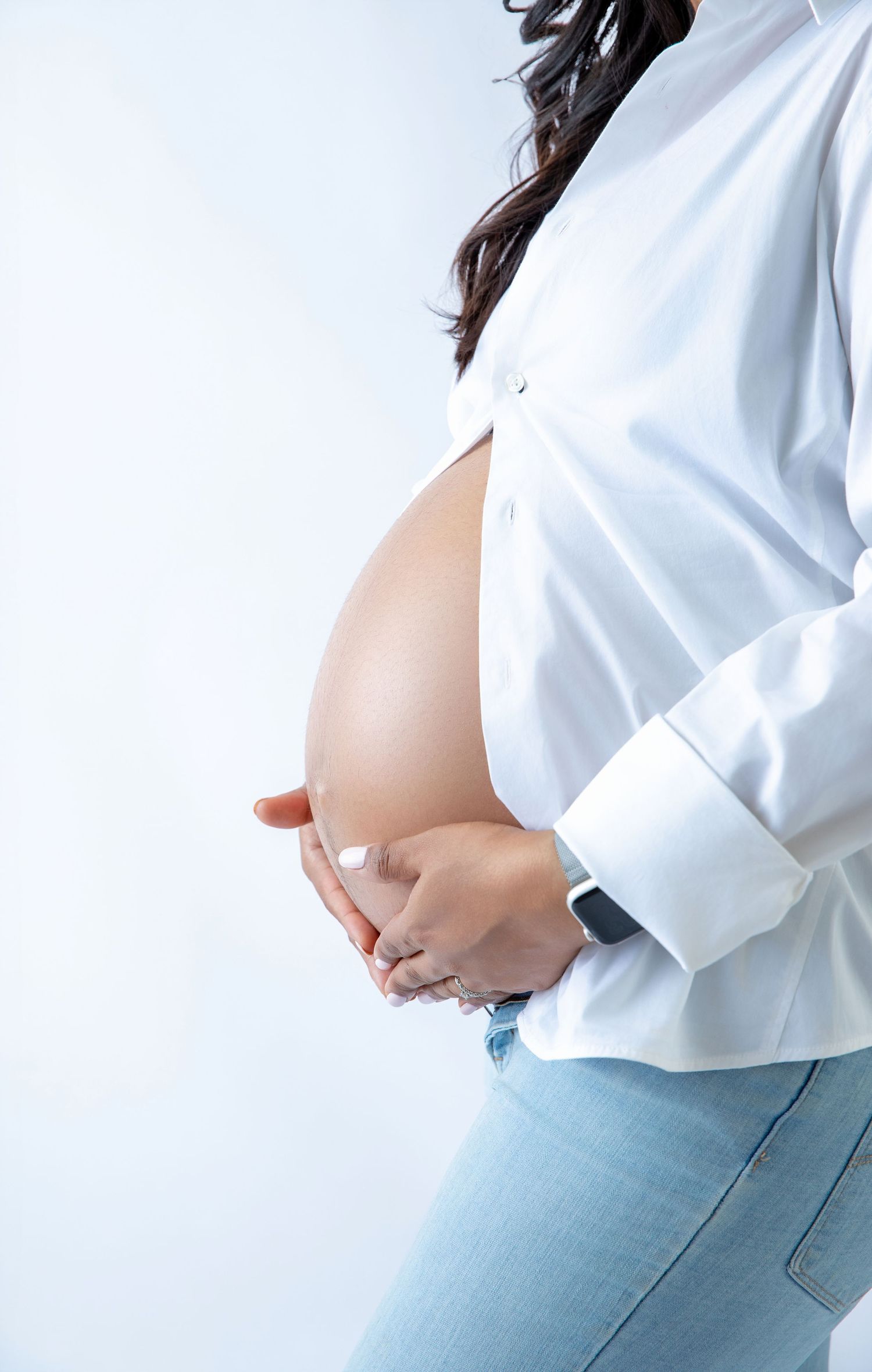 Close-up of pregnant woman in open white shirt and jeans gently holding bare baby bump.