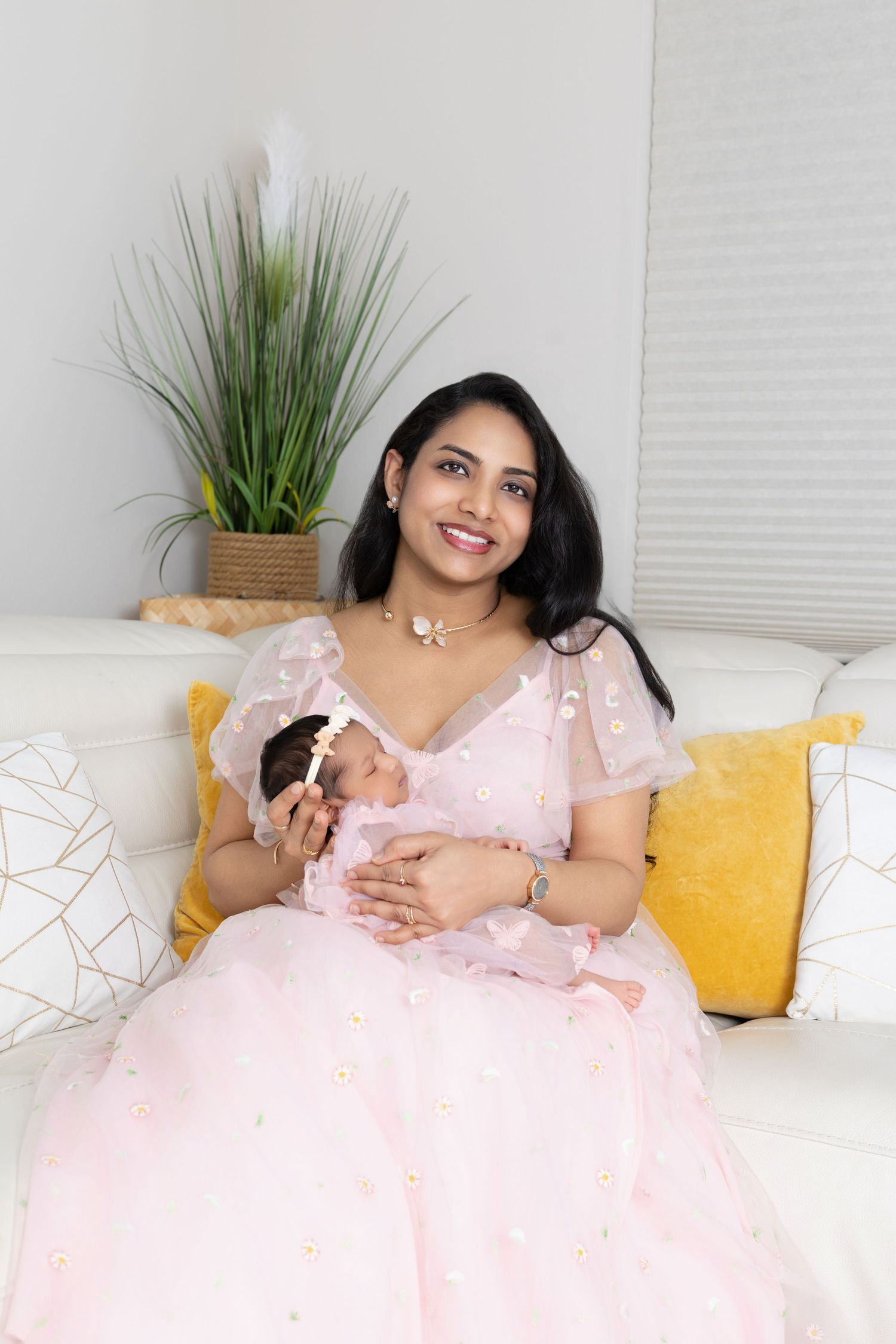 Smiling Indian mother in pink dress holds sleeping newborn baby on white sofa with yellow pillows.