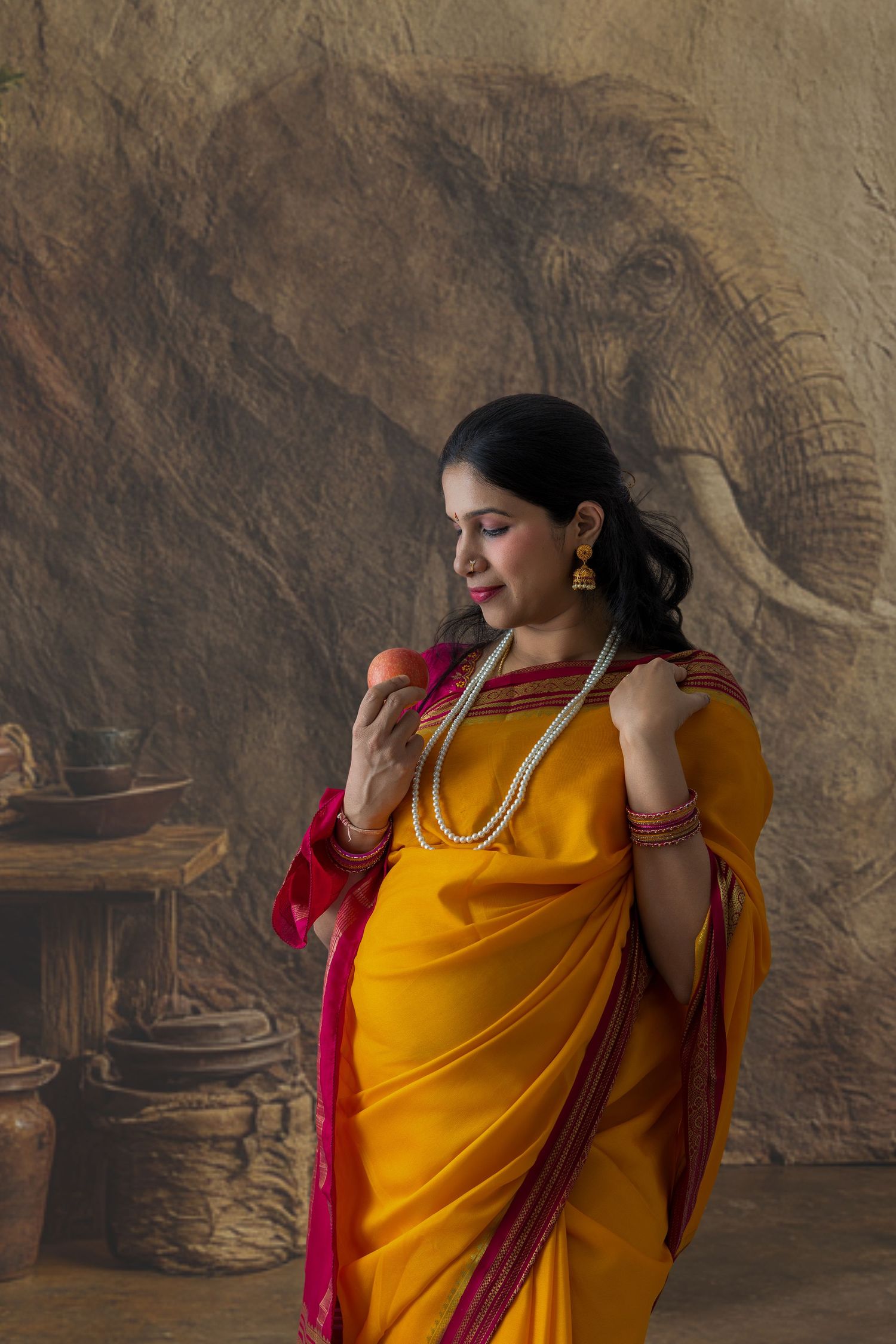 Woman in yellow and red saree holding fruit, pearl necklace, elephant mural backdrop, traditional Indian attire.