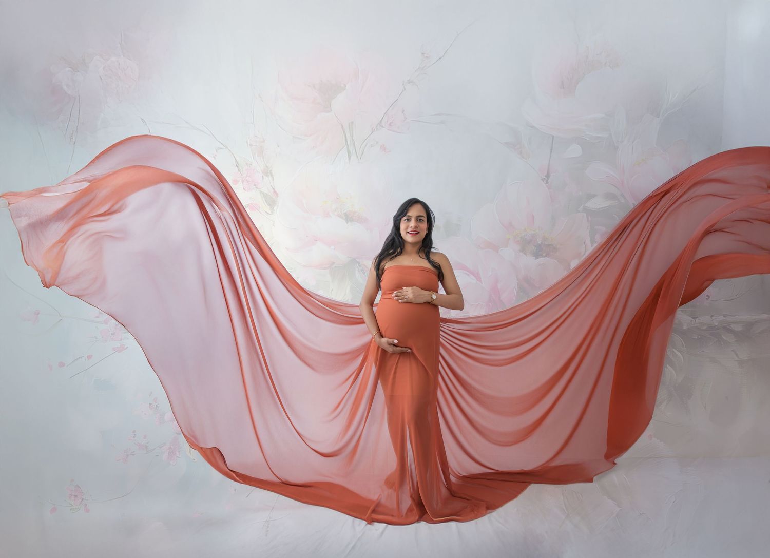 Pregnant woman in flowing coral maternity gown with dramatic fabric wings against soft floral backdrop.