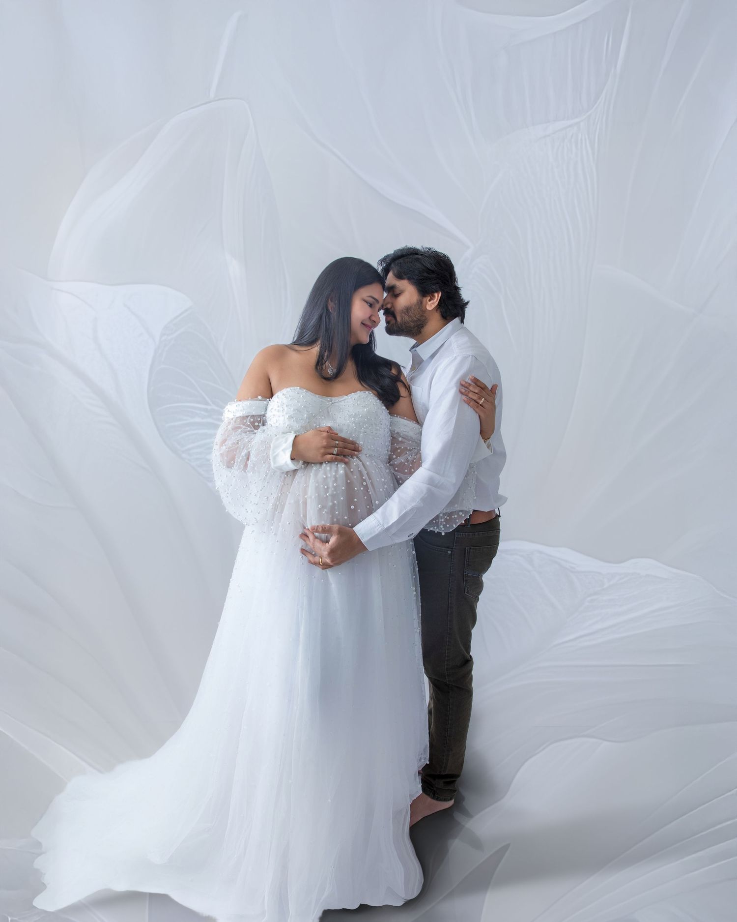 Romantic maternity photo session with couple in white outfits against ethereal flowing fabric backdrop.