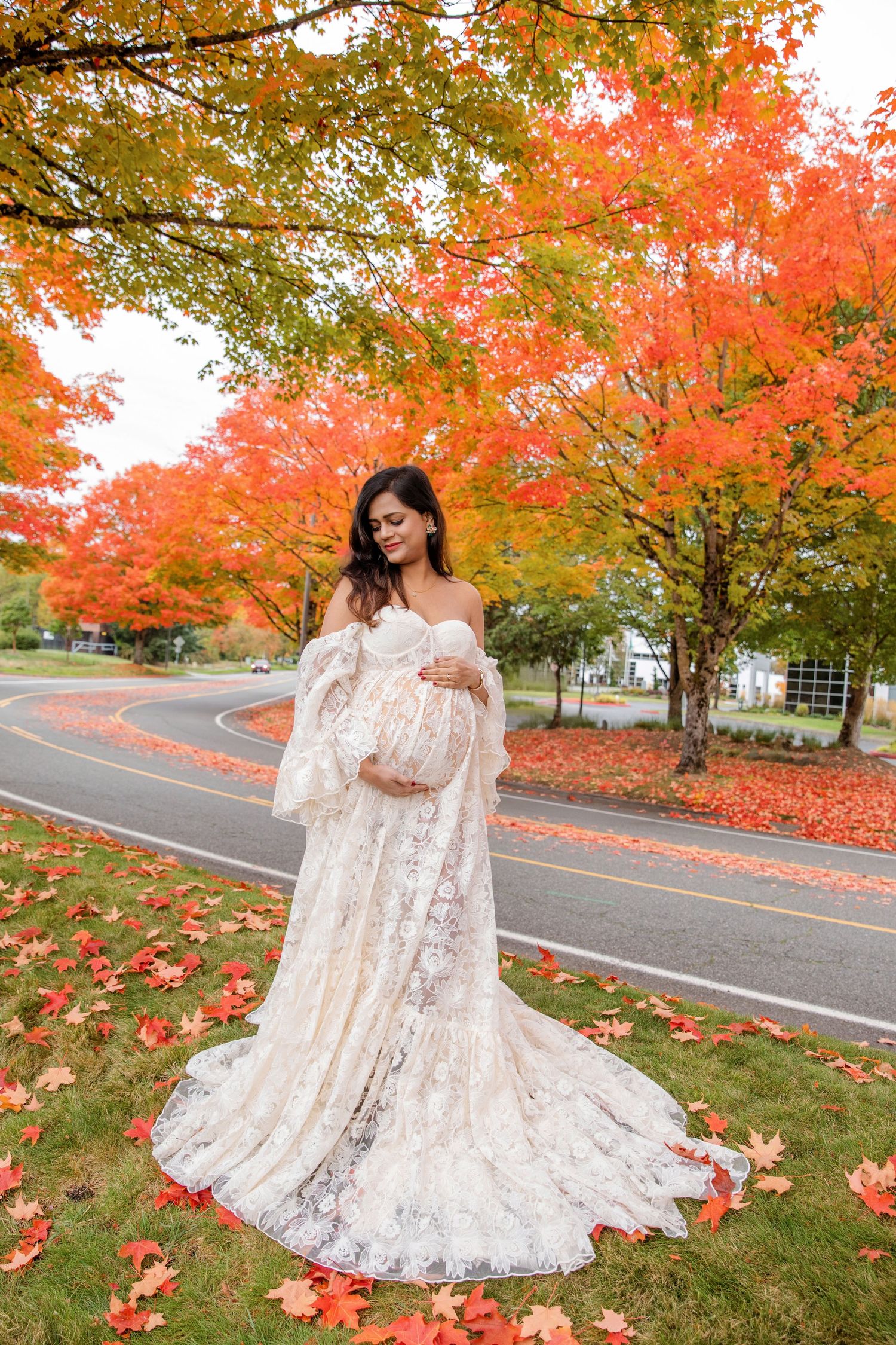 A lace maternity gown photoshoot amidst vibrant fall foliage with red maple trees and scattered autumn leaves.