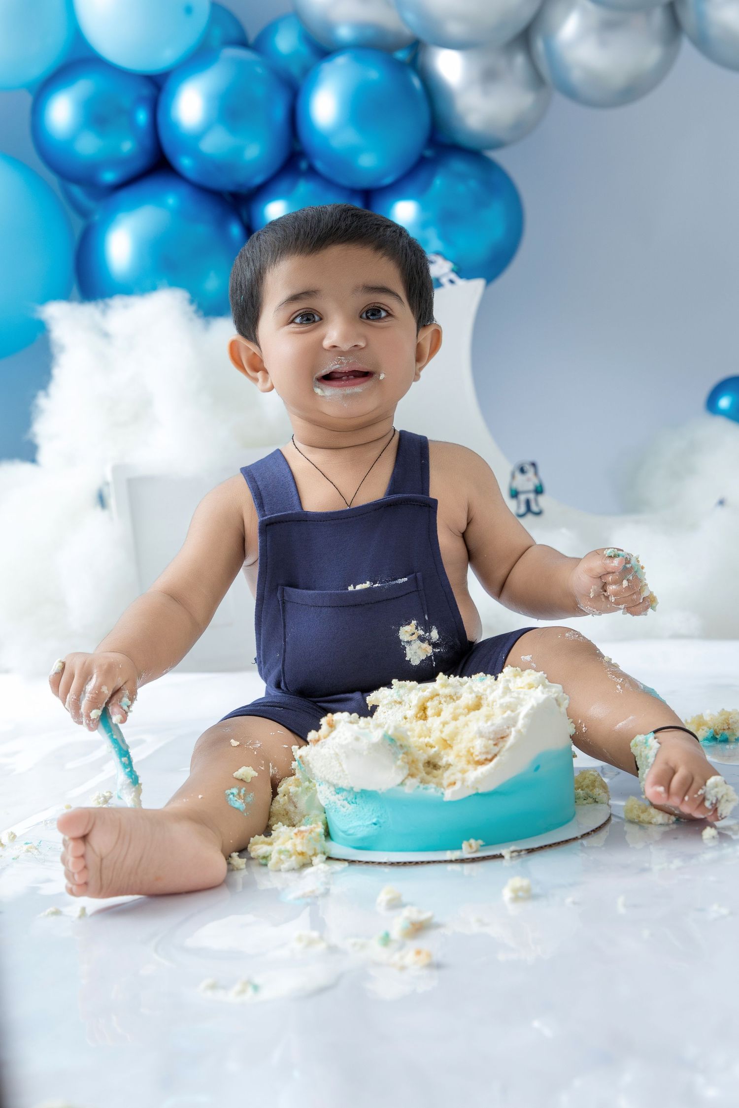 Baby playing with cake during first birthday photoshoot surrounded by blue and silver balloons.