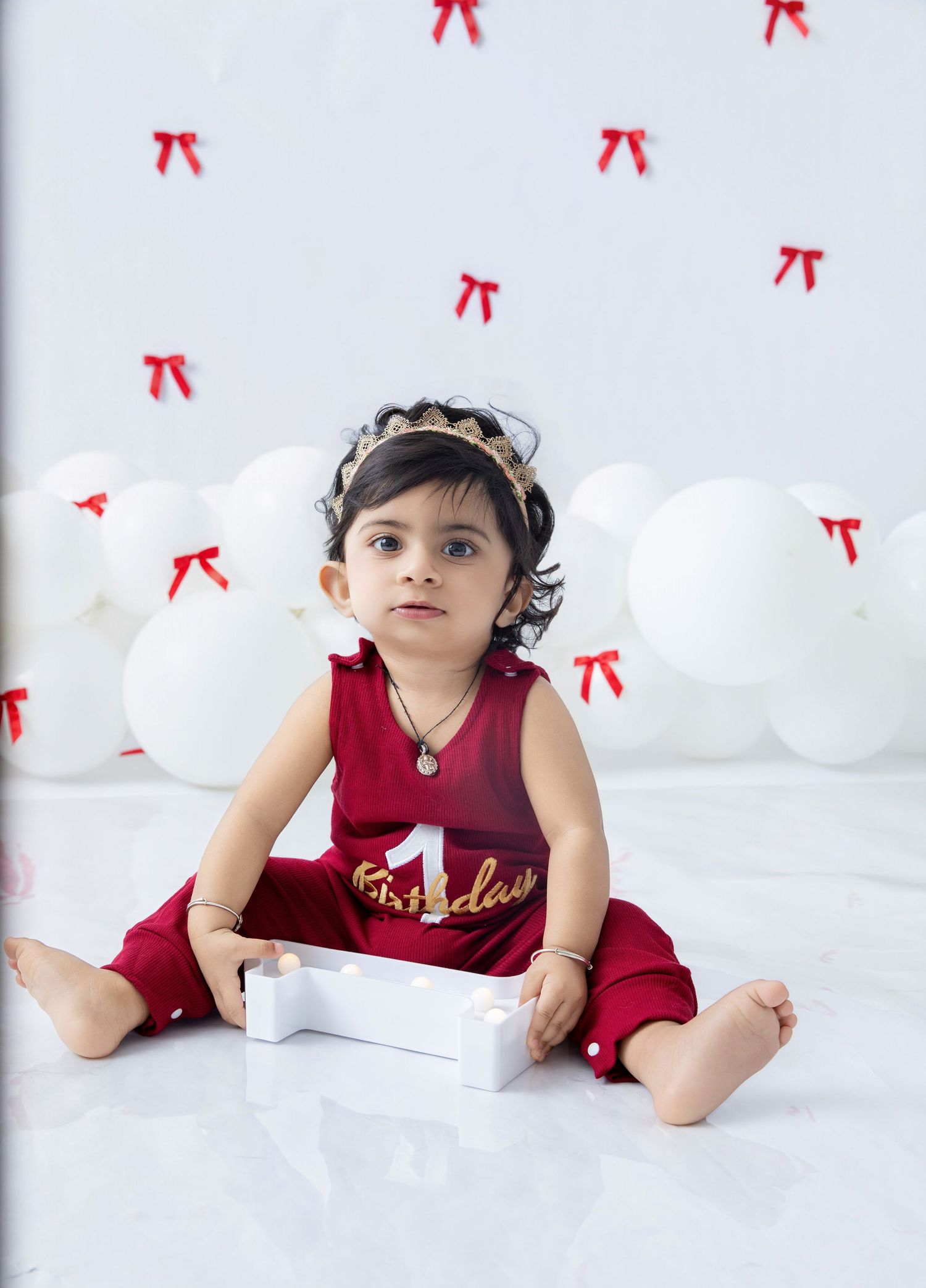 A baby in a burgundy outfit lies on a white backdrop with red bows scattered around the scene.