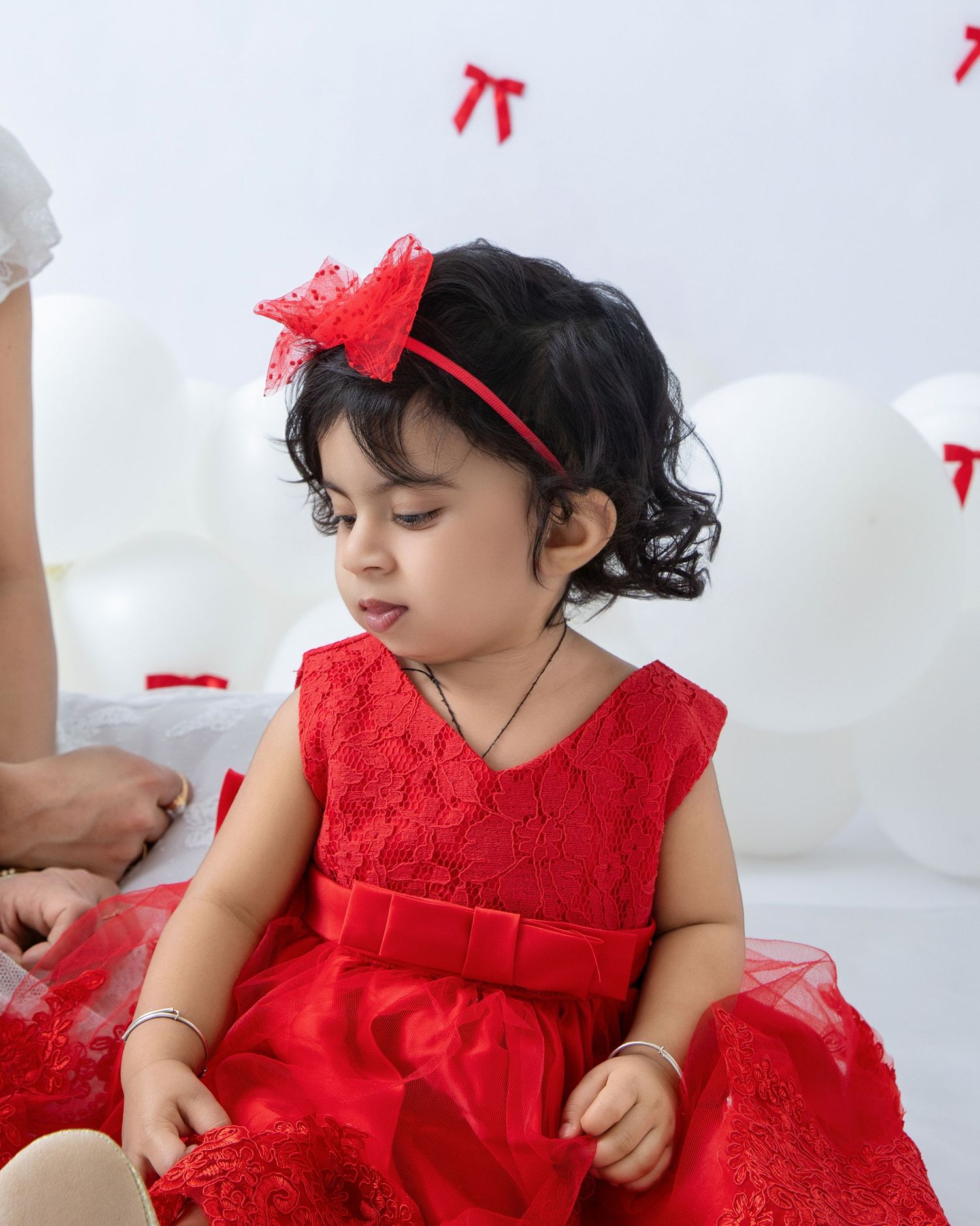 A toddler wearing a festive red dress poses against a white backdrop with red bow decorations.