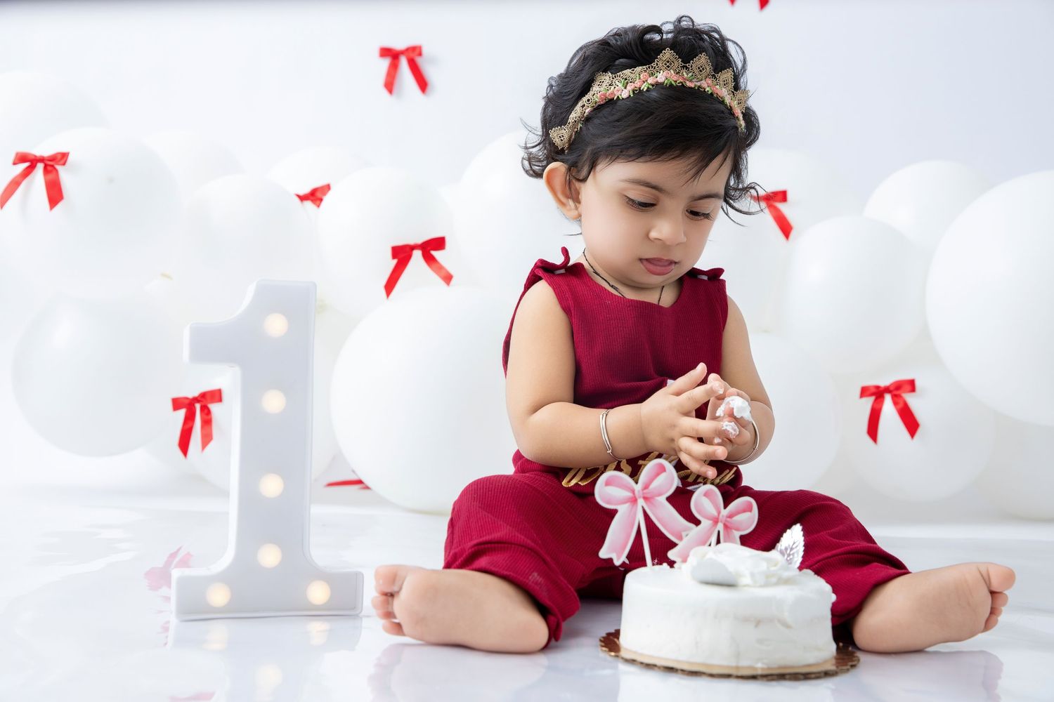 Baby in red outfit touching white cake during first birthday photoshoot.