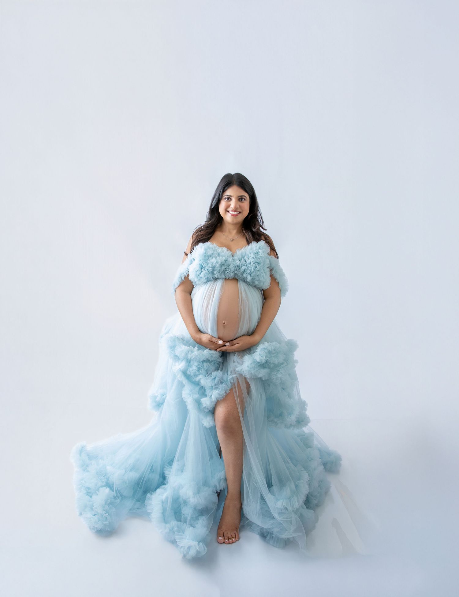 Pregnant woman in a flowing light blue ruffled tulle maternity gown posing gracefully against a white background.