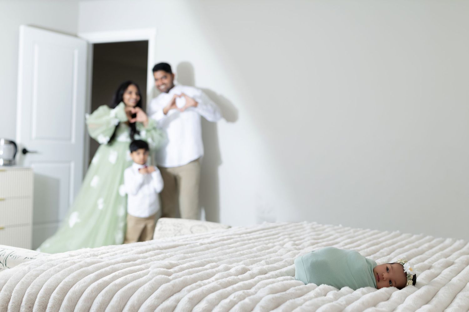 Newborn baby sleeping on a bed while happy family with young child stands in the background in a bright bedroom.