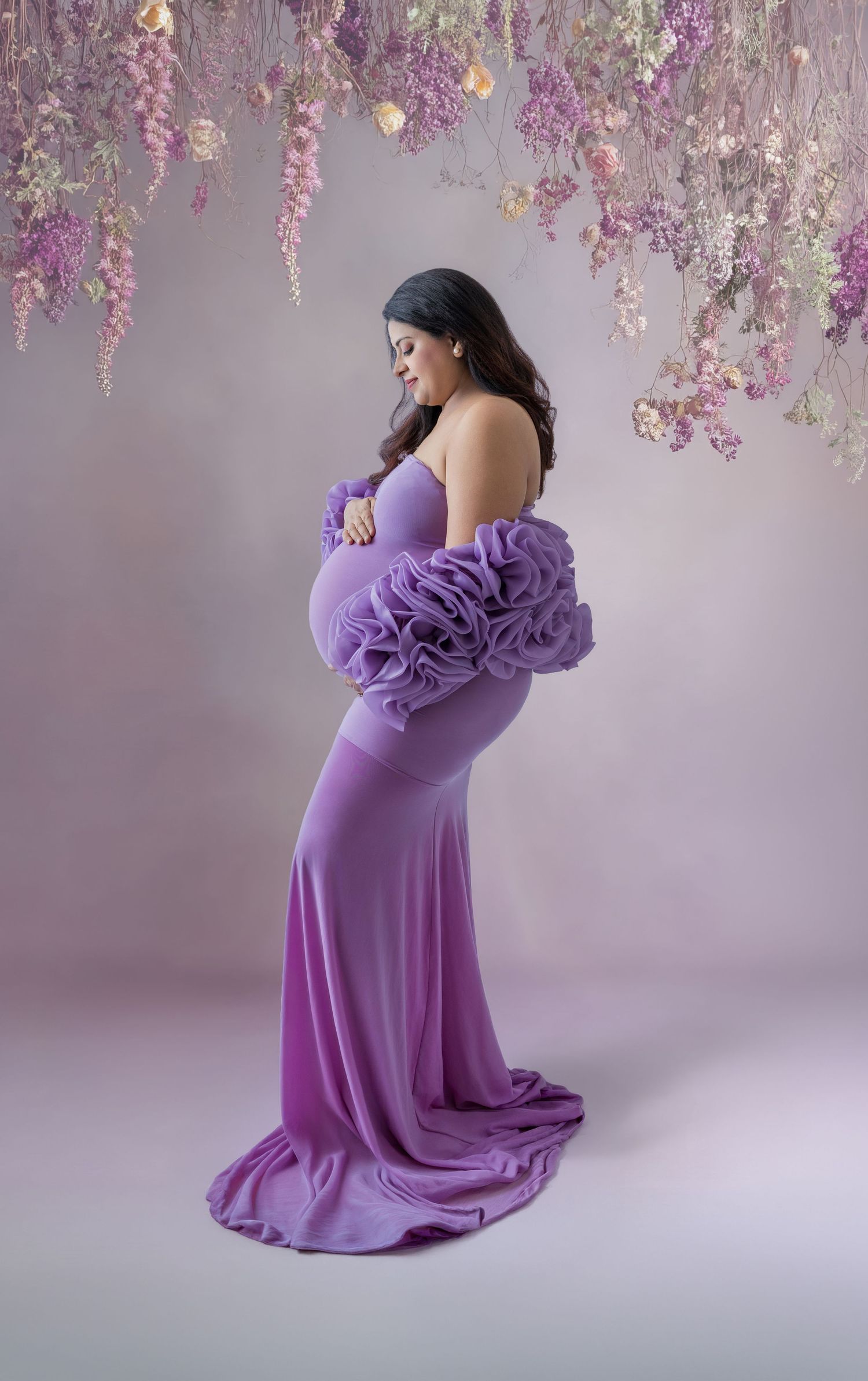 Pregnant woman in purple ruffle gown poses gracefully under hanging floral arrangement in a studio maternity photo.