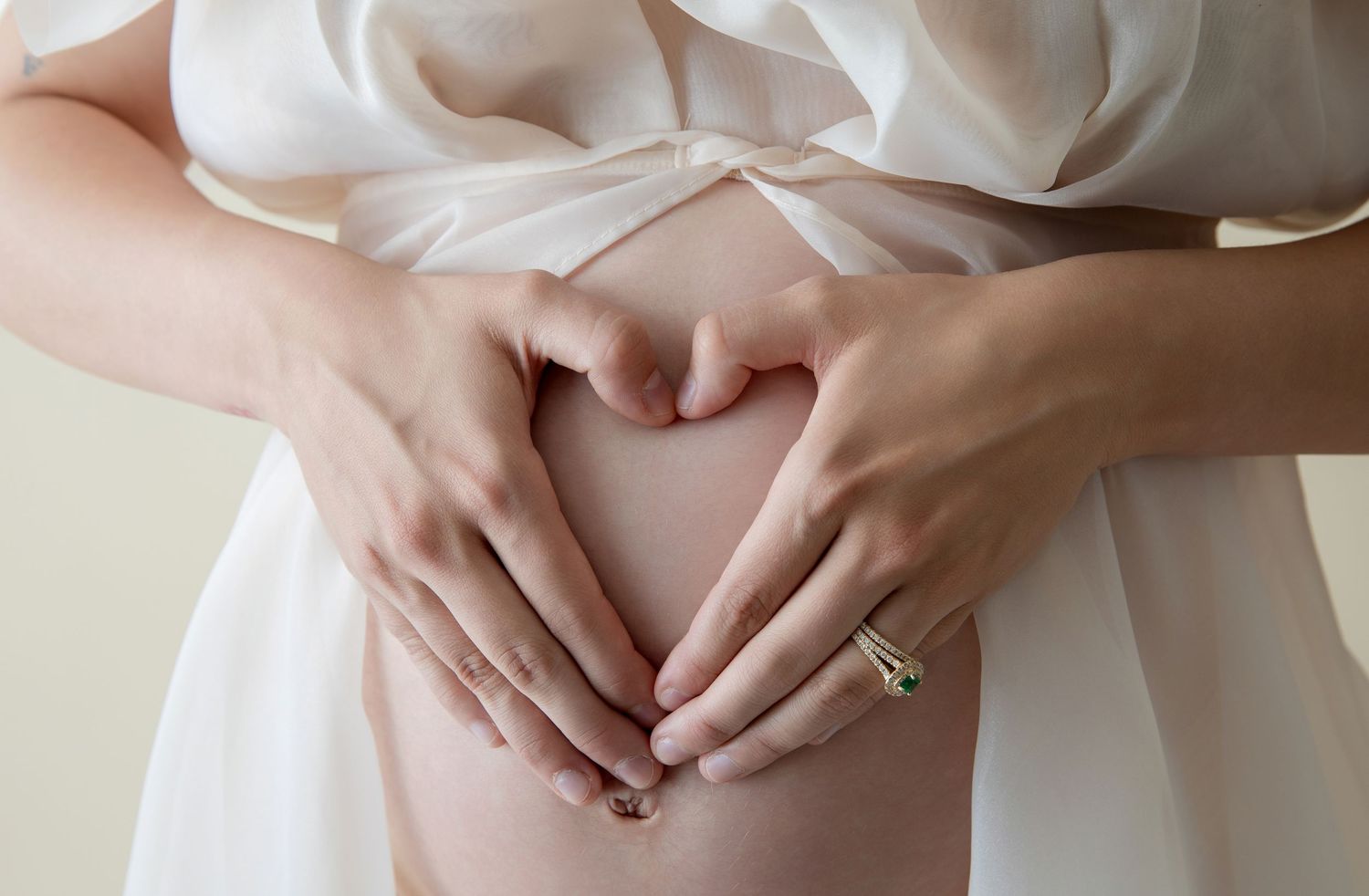 Pregnant woman forming a heart shape with her hands on her baby bump, wearing a white dress and ring.