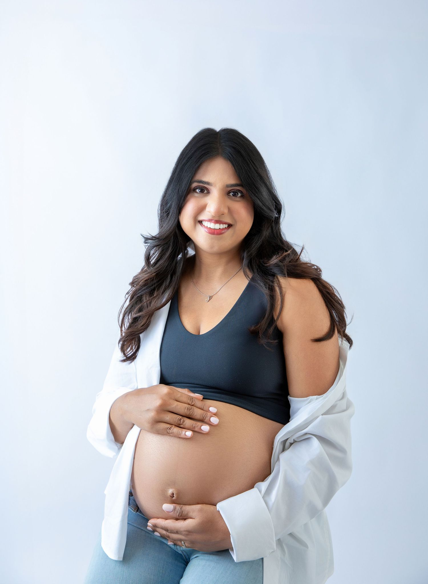 Smiling pregnant woman in dark sports bra and white blazer holds baby bump against light background.