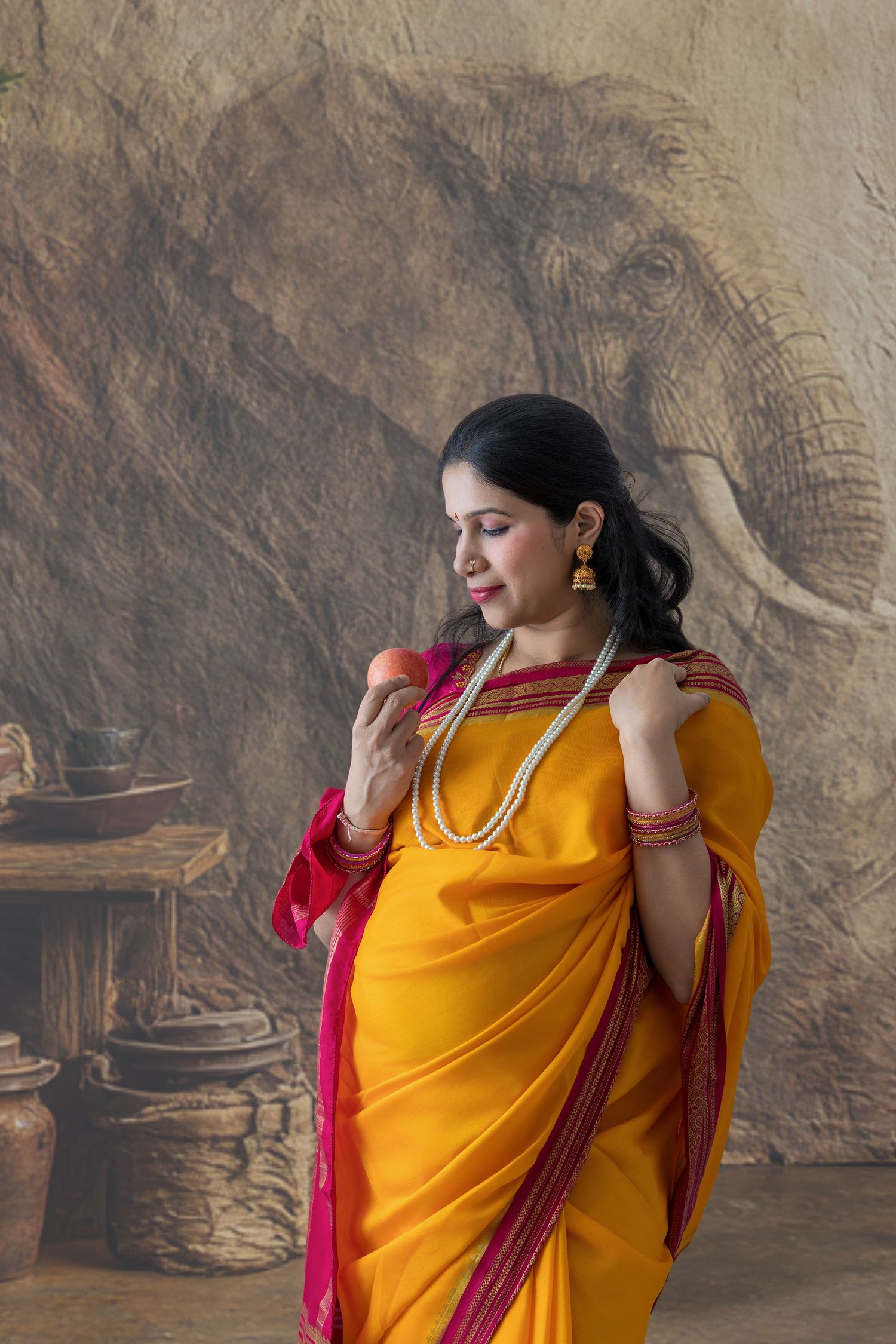 Pregnant woman in vibrant yellow and pink saree with pearl necklace posing before elephant mural backdrop.
