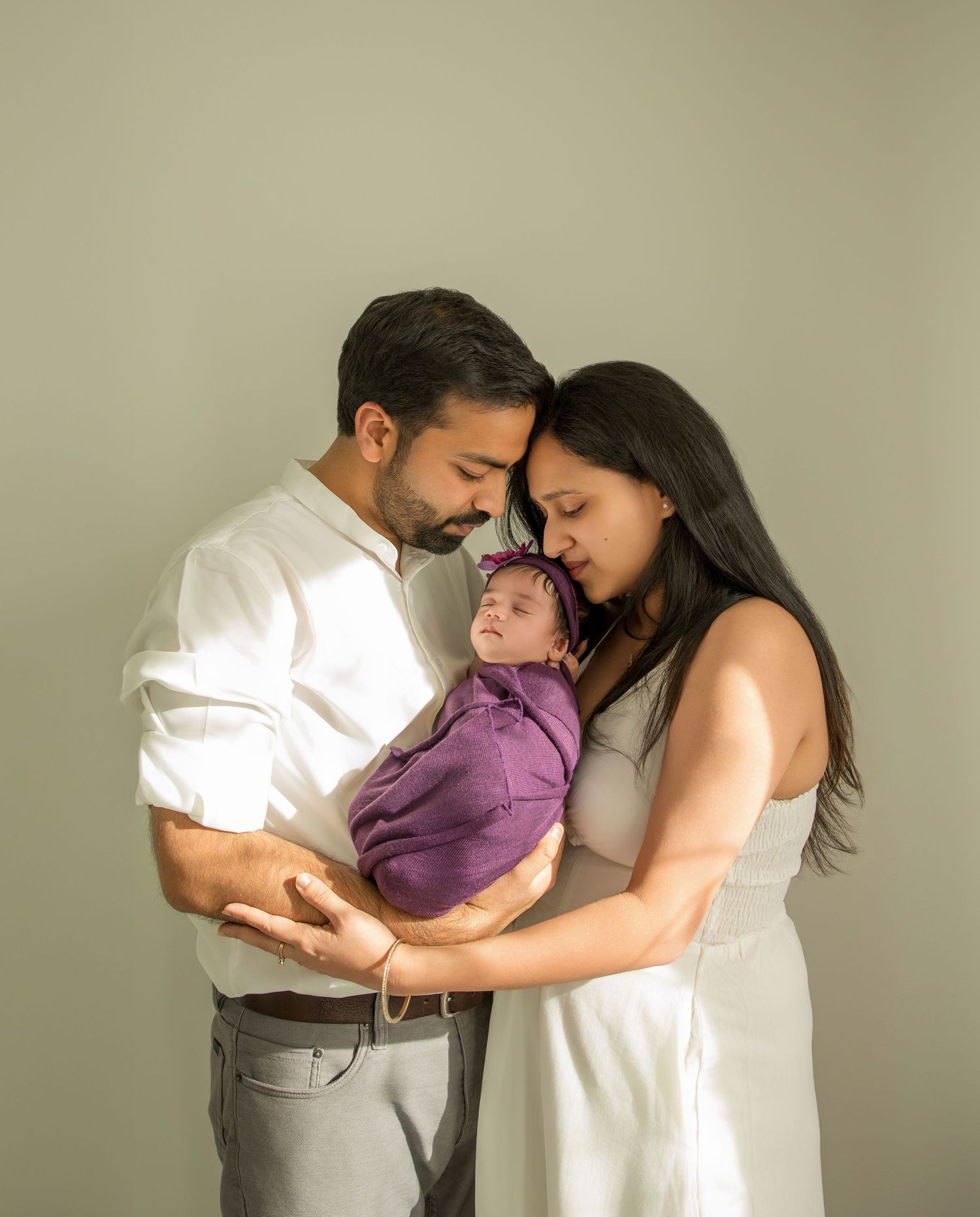 A couple in white outfits share an intimate moment with their baby wearing purple against a neutral background.
