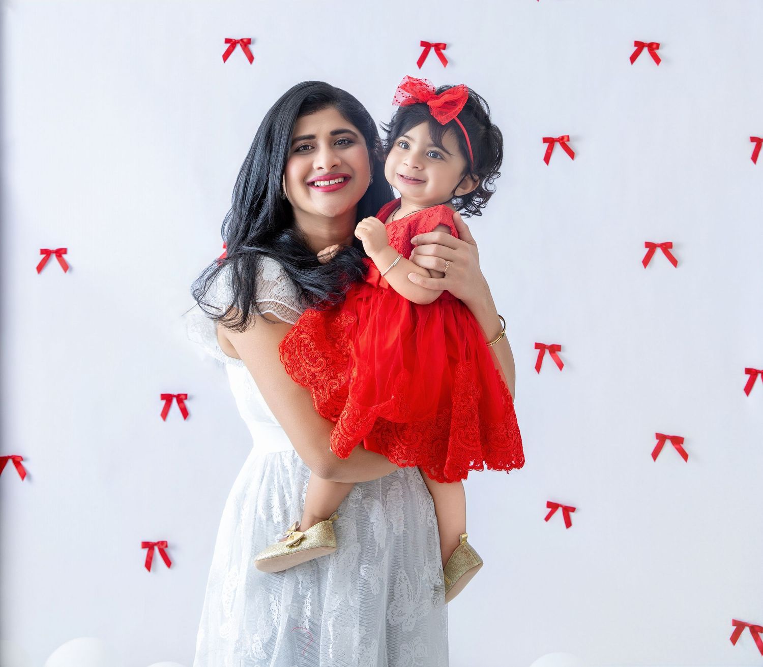 Mother and child in matching white and red dresses pose against wall decorated with red bows.