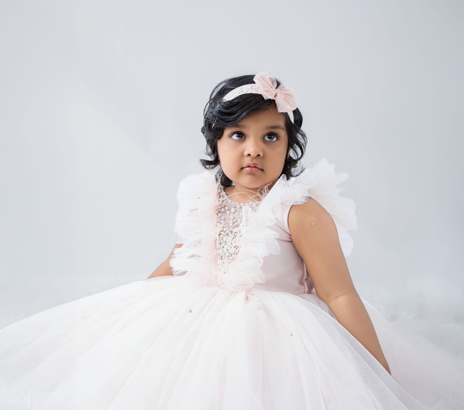 Toddler wearing an elegant white tulle dress and pink hair bow against a light gray background.