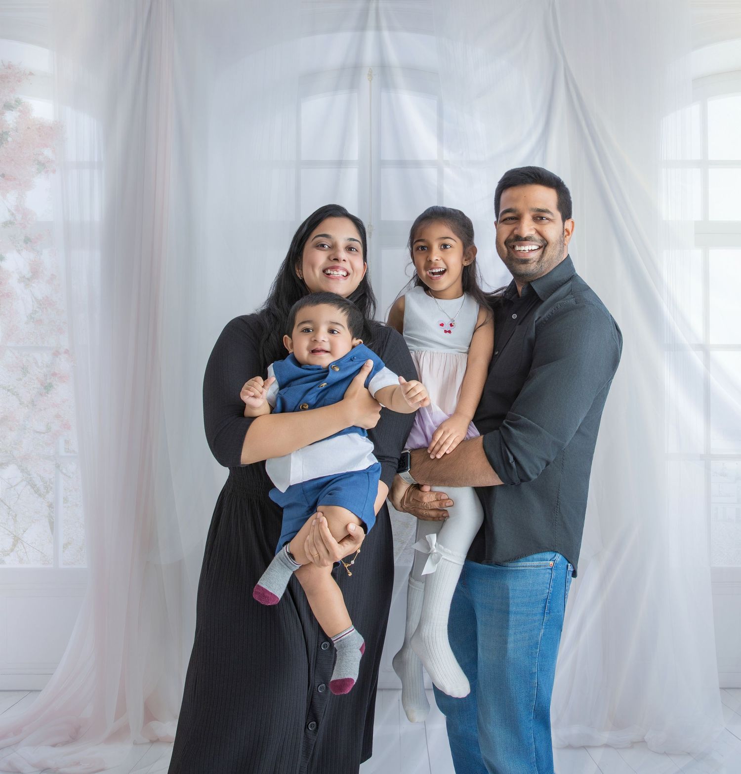 A family poses together in a bright, airy room with white curtains and natural lighting.