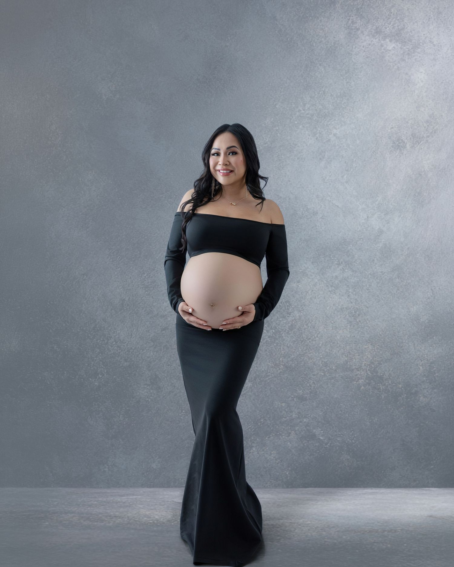 Pregnant woman in black off-shoulder crop top and maxi skirt cradling baby bump during maternity photo session.