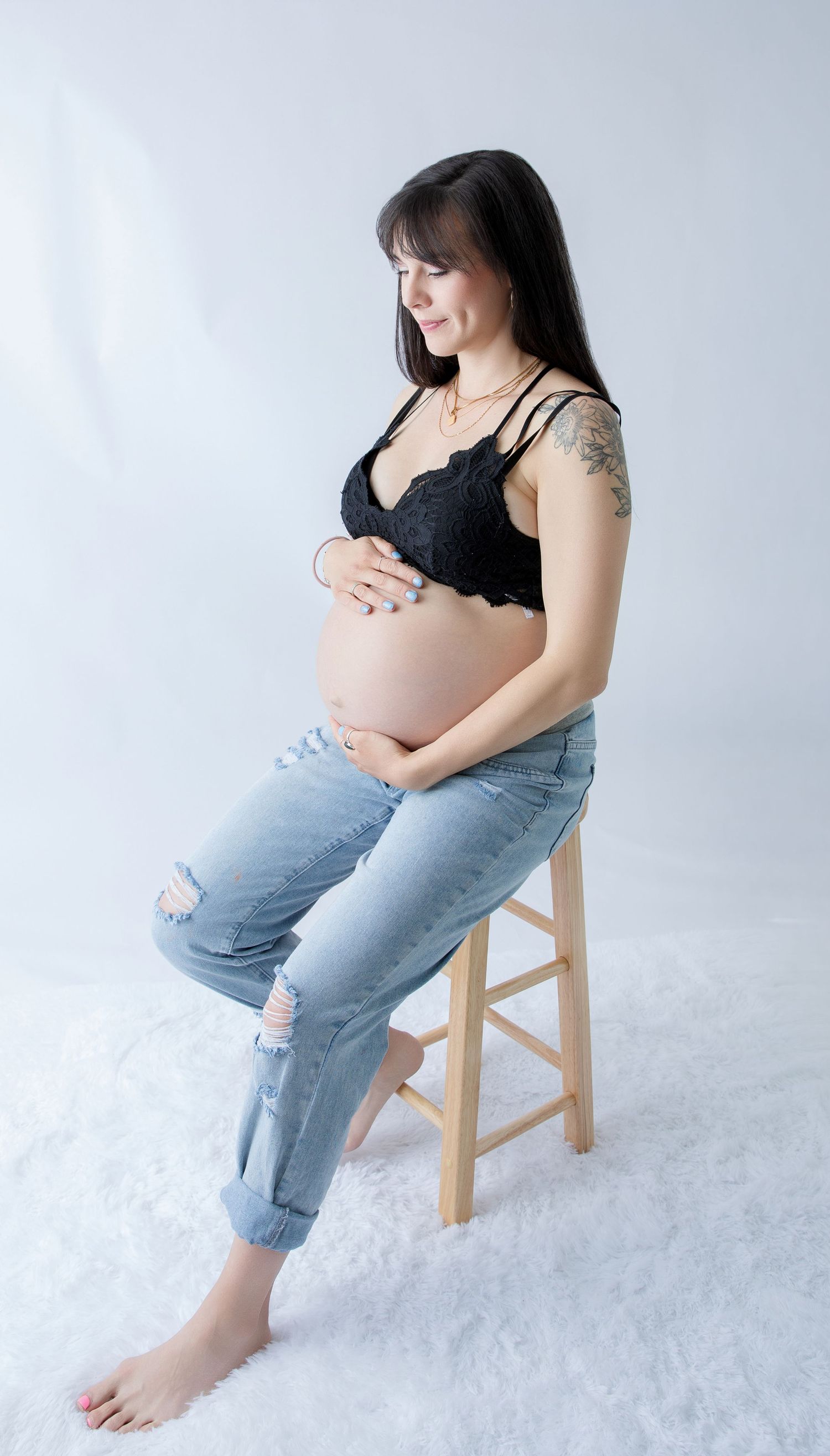 Pregnant woman in black lace bralette and ripped jeans sits on stool cradling baby bump in studio.