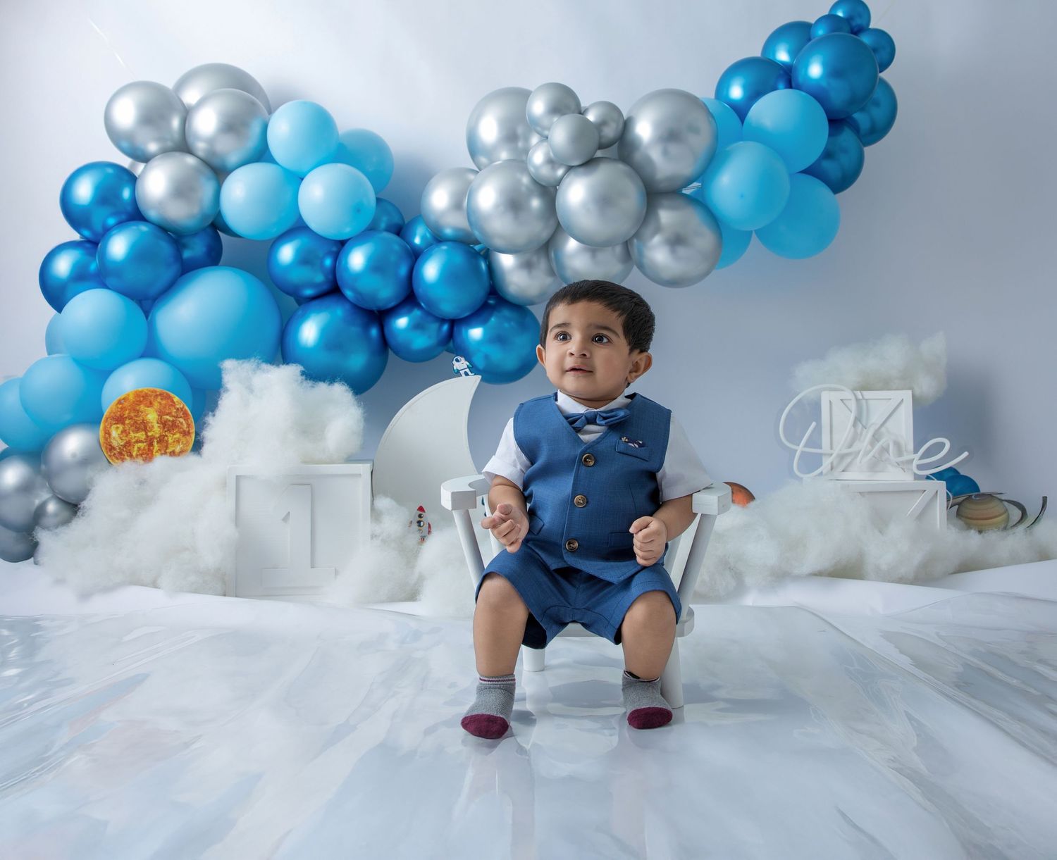 A child sits in a white studio with blue and silver balloon decorations and cloud-like elements in the background.