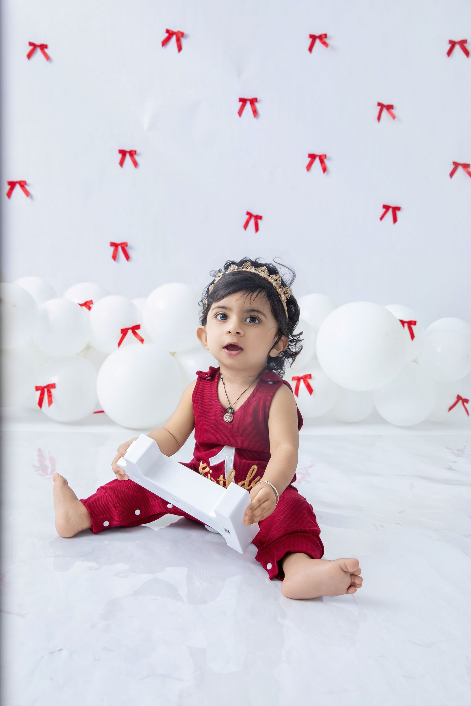 Baby in red dress lying on white floor with decorative bows and number one light.