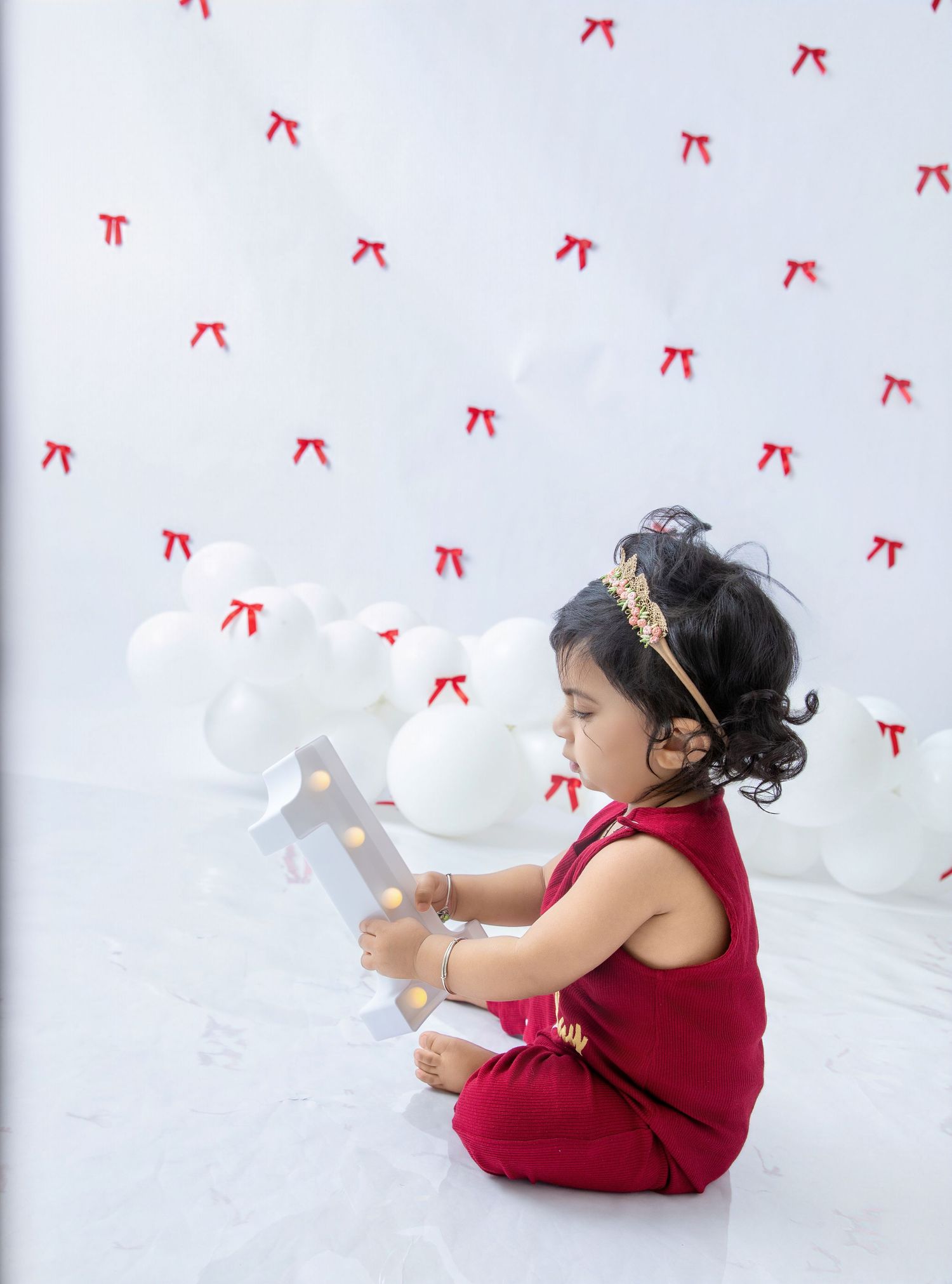 A toddler in red clothing plays with white balloons against a backdrop decorated with red bows.