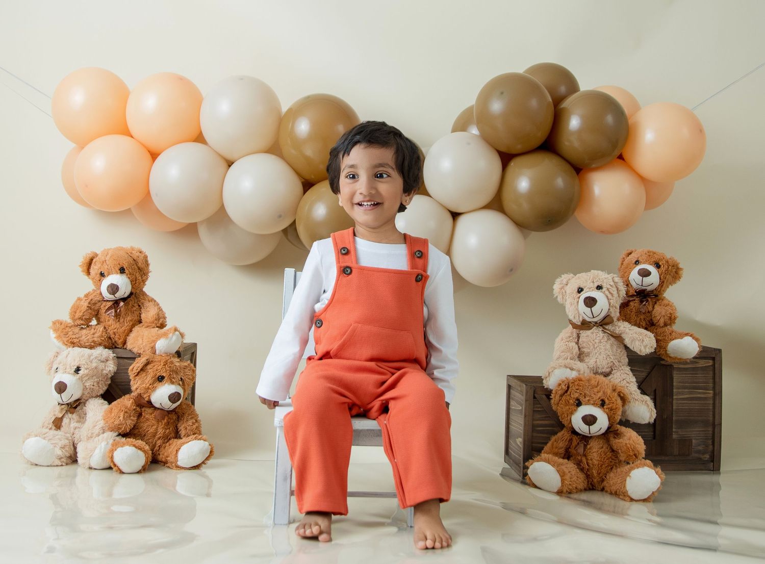 Child in coral overalls sits on wooden crate with teddy bears and balloons in background.