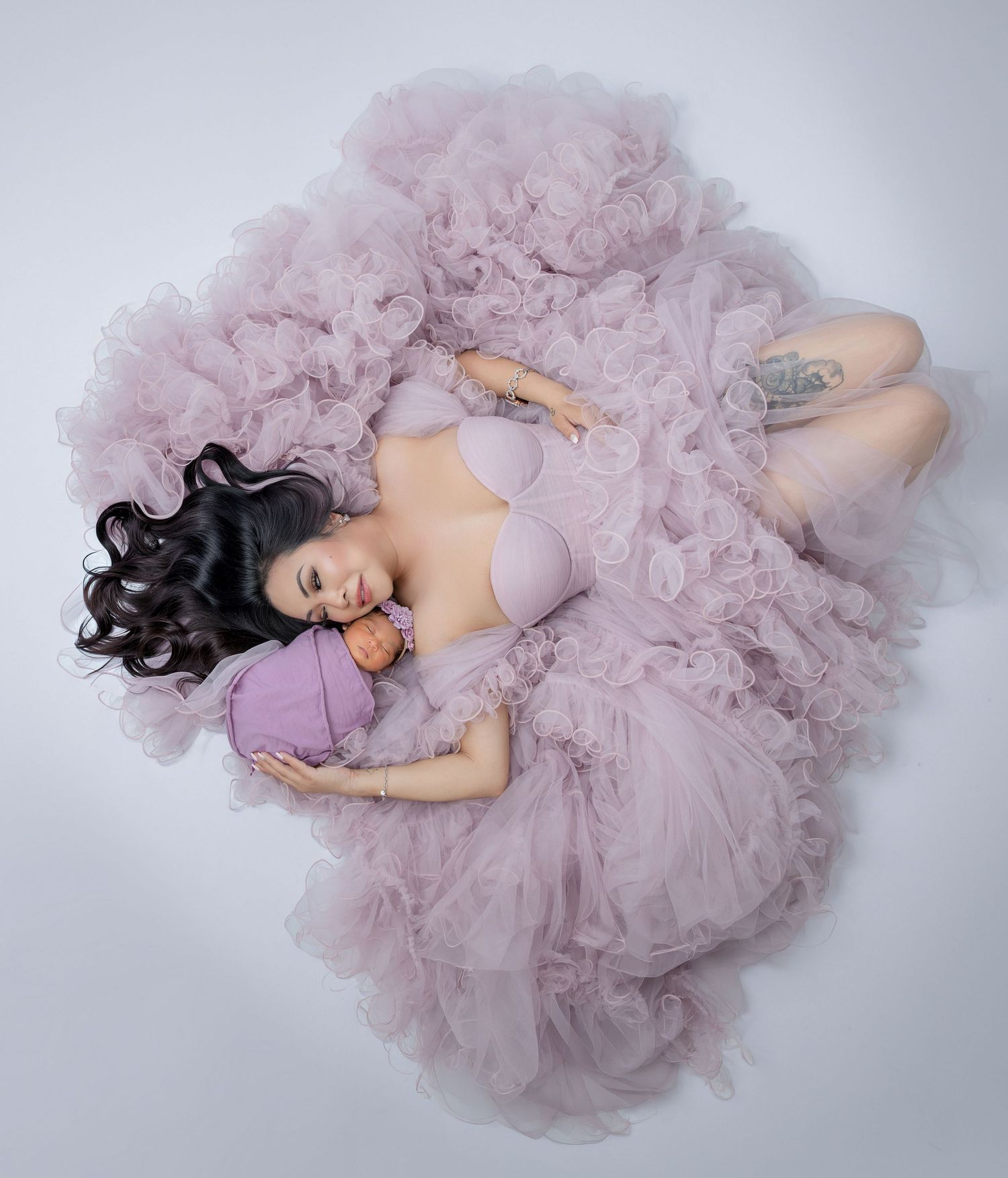 Mother in lavender tulle gown lying down cradling swaddled newborn baby from above.