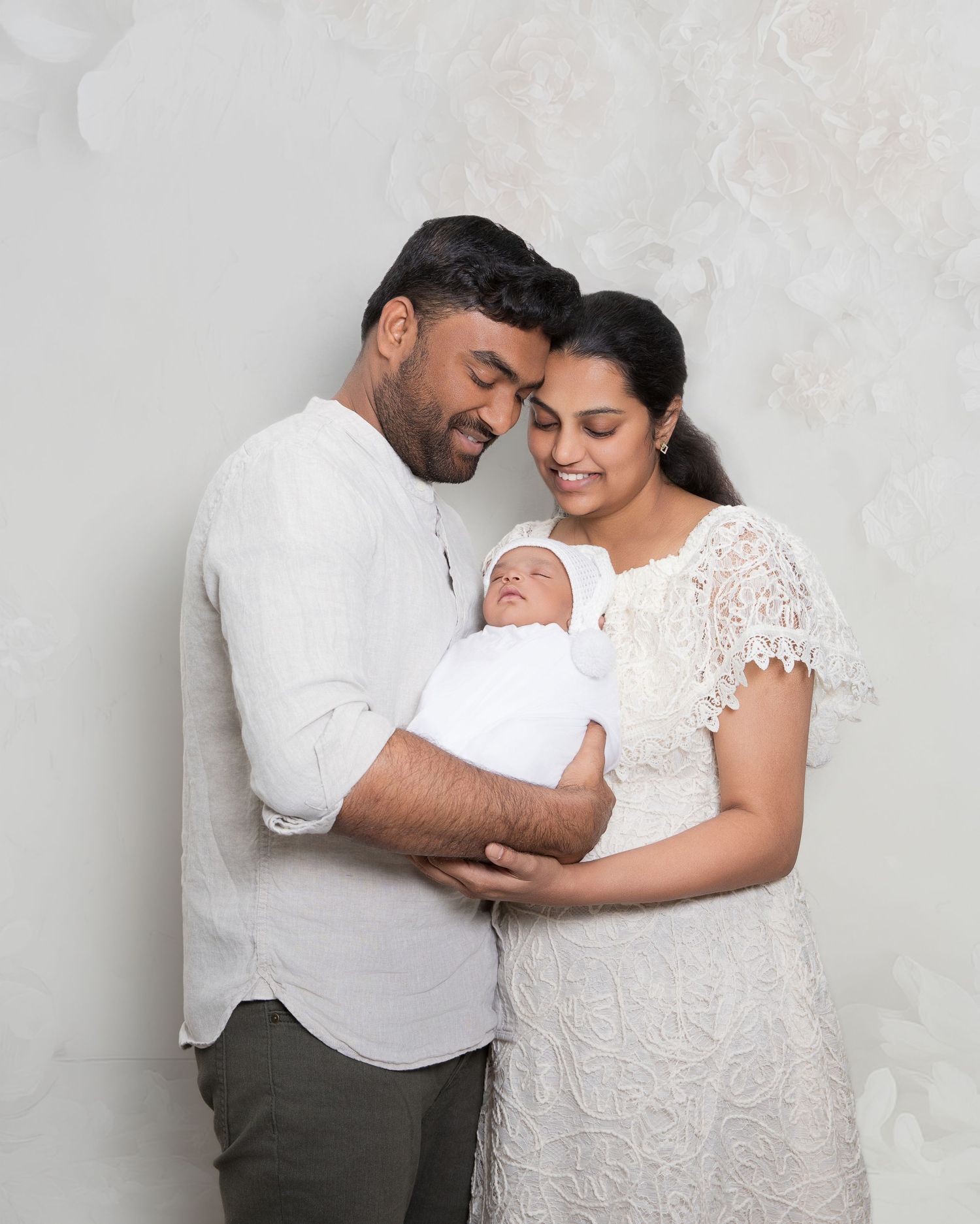 New parents in white outfits tenderly hold their swaddled newborn baby against a floral white backdrop.