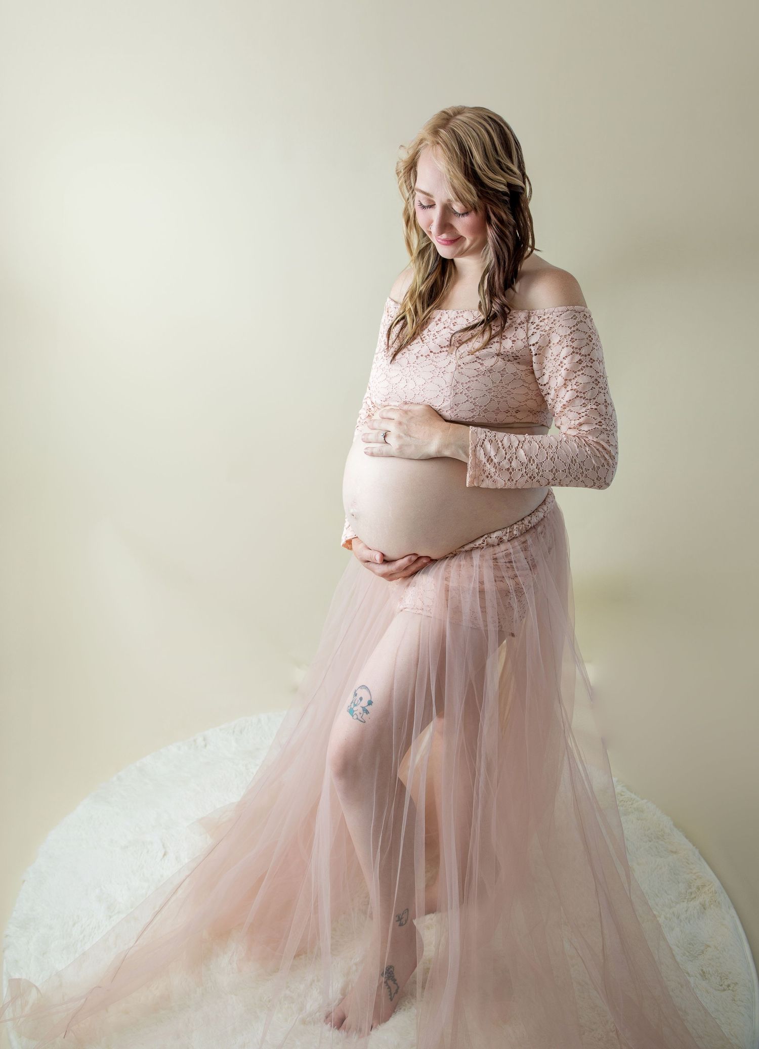 Pregnant woman in blush pink lace and tulle maternity gown cradling her baby bump in a soft studio portrait.