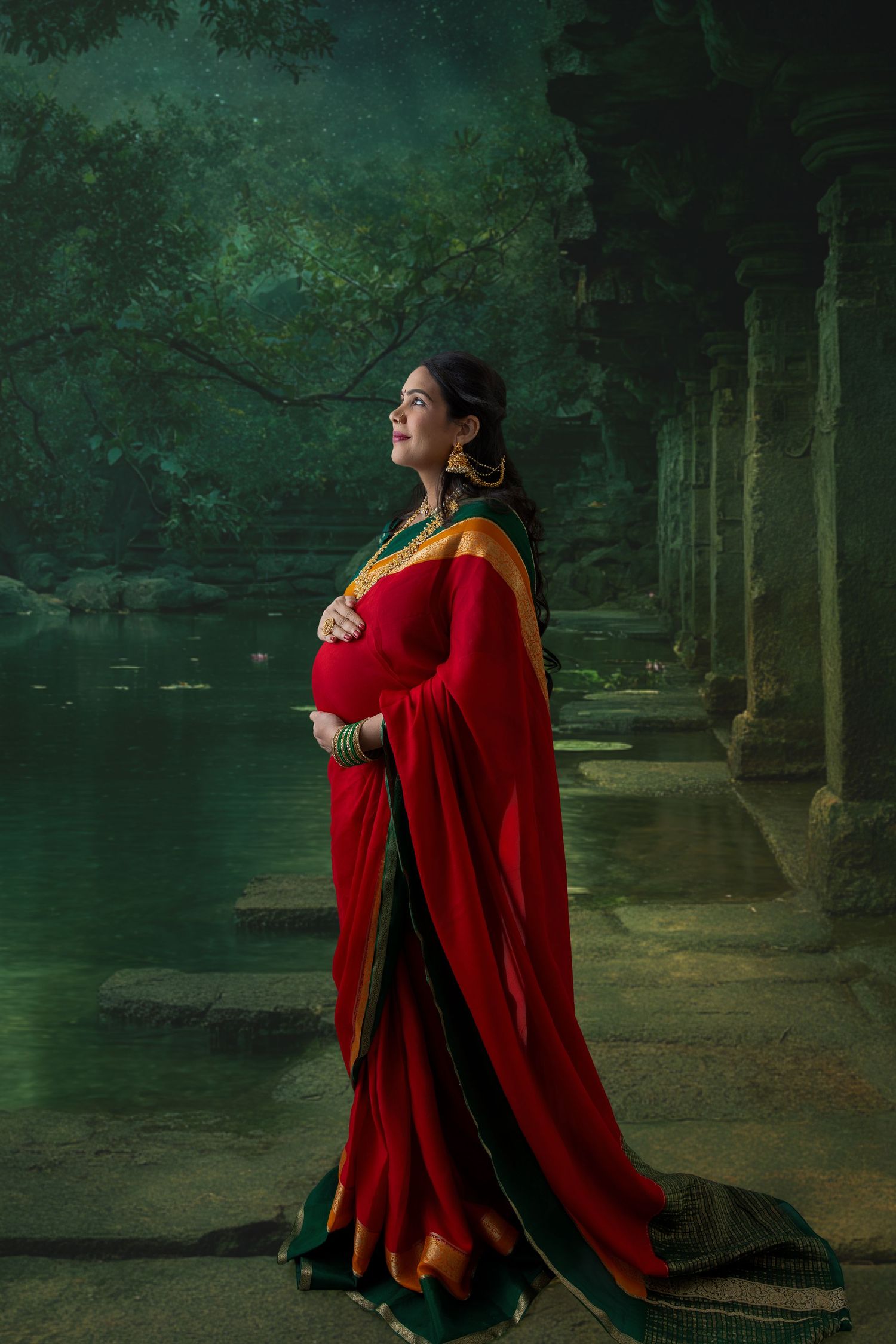 A pregnant woman in a red and green saree stands gracefully by a serene forest pool near ancient stone columns.