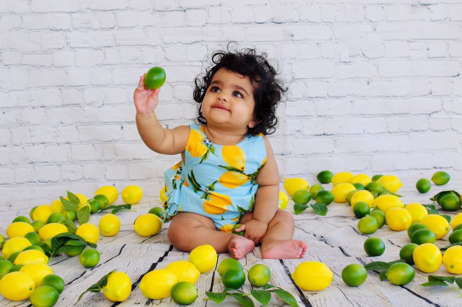 Baby in blue dress lying on white floor with scattered yellow lemons and green limes.