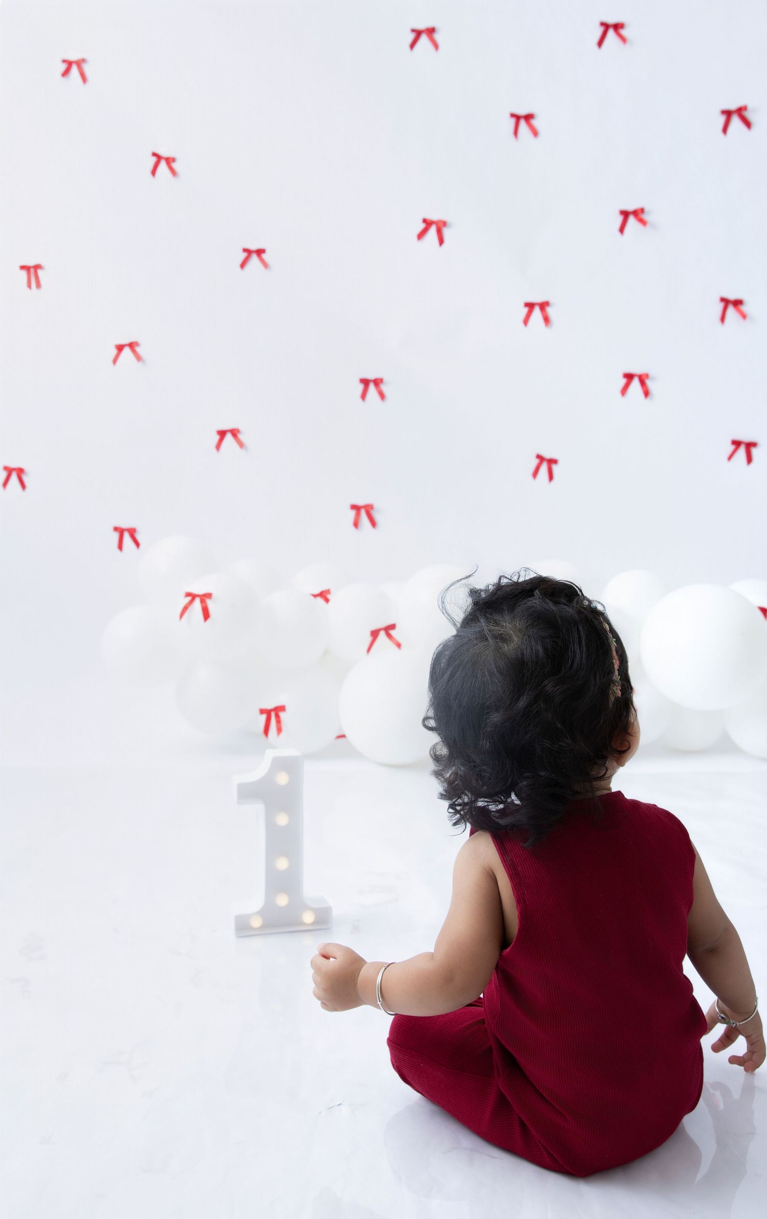 Baby in red outfit sits facing white number one decoration against bow-patterned backdrop.