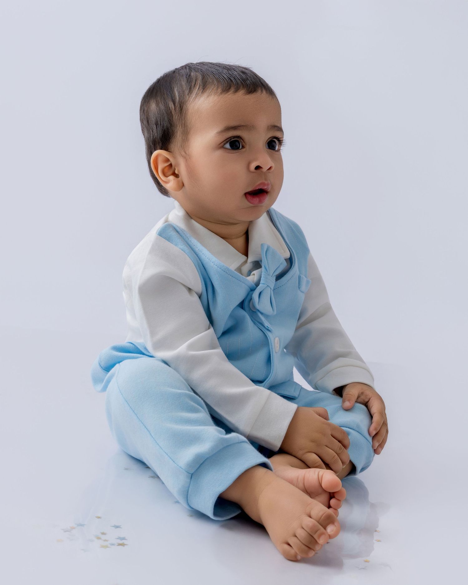 A small child in a light blue overalls and white long sleeve shirt sits on a white background looking to the side.