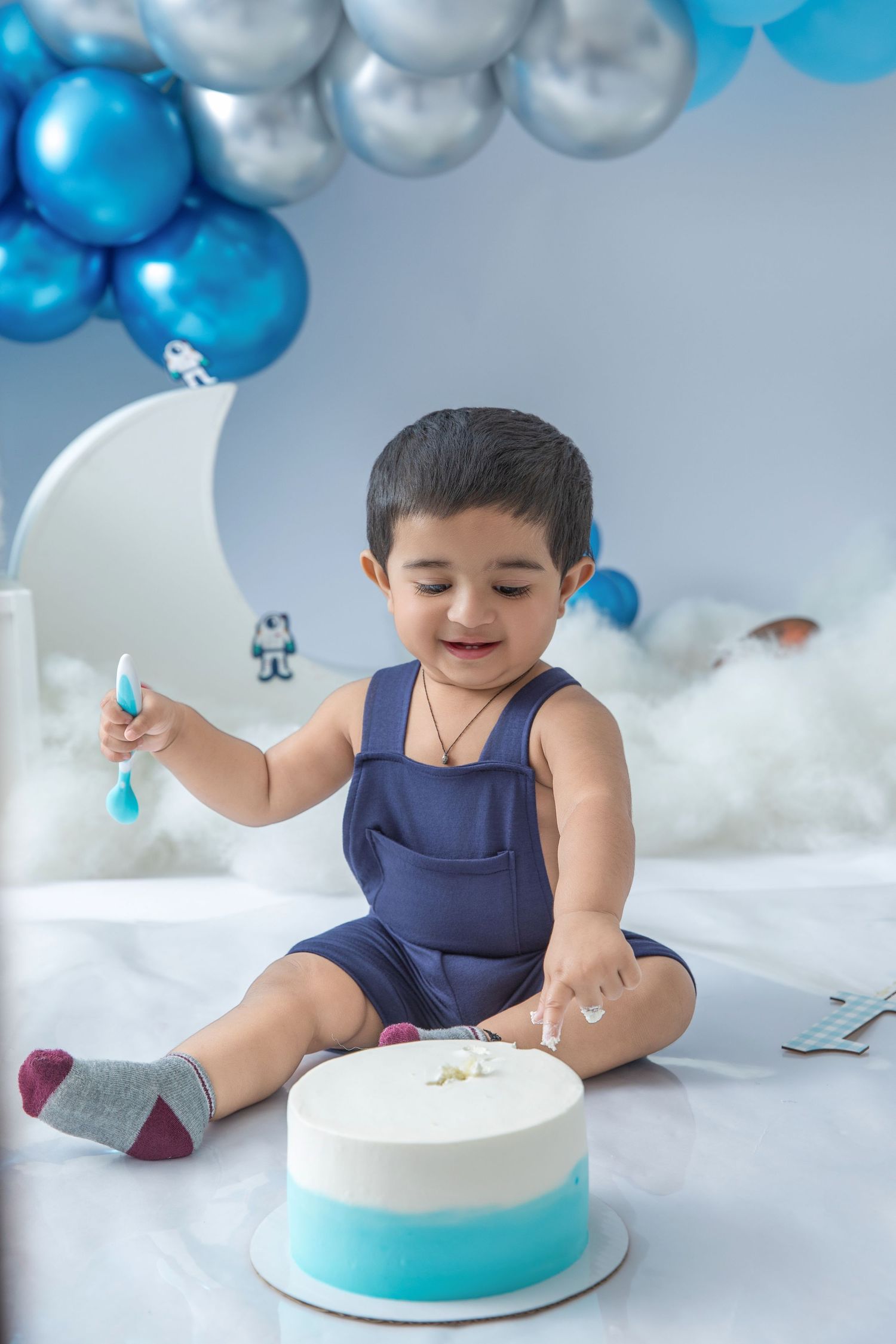 A cake smash photo setup with blue and silver balloons, white clouds, and a turquoise ombre cake.