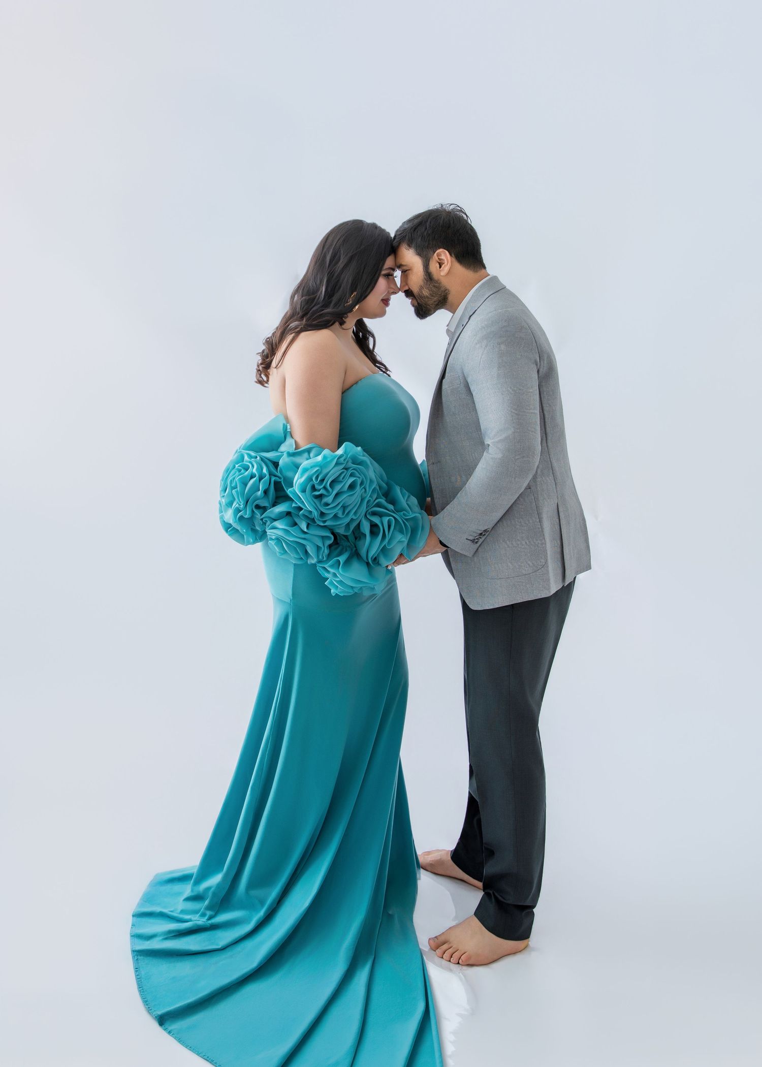 Couple in maternity photo shoot, woman in teal gown with floral sleeves, man in gray suit, foreheads touching.