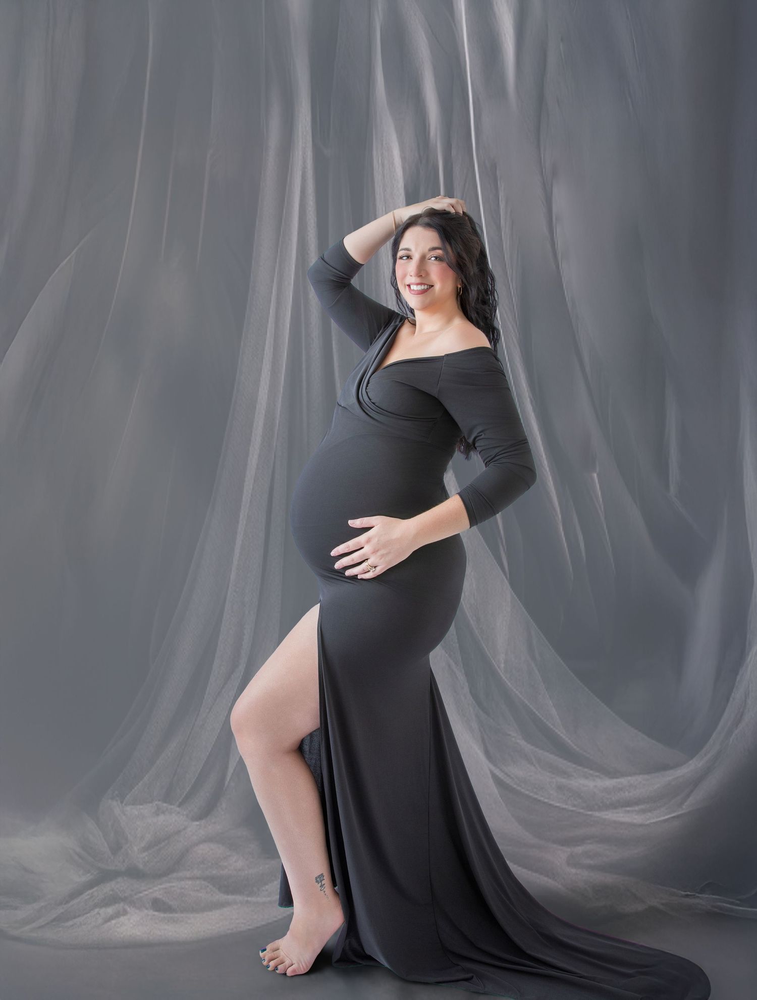 Pregnant woman in elegant dark gray off-shoulder maternity gown poses gracefully against sheer silver draped backdrop.