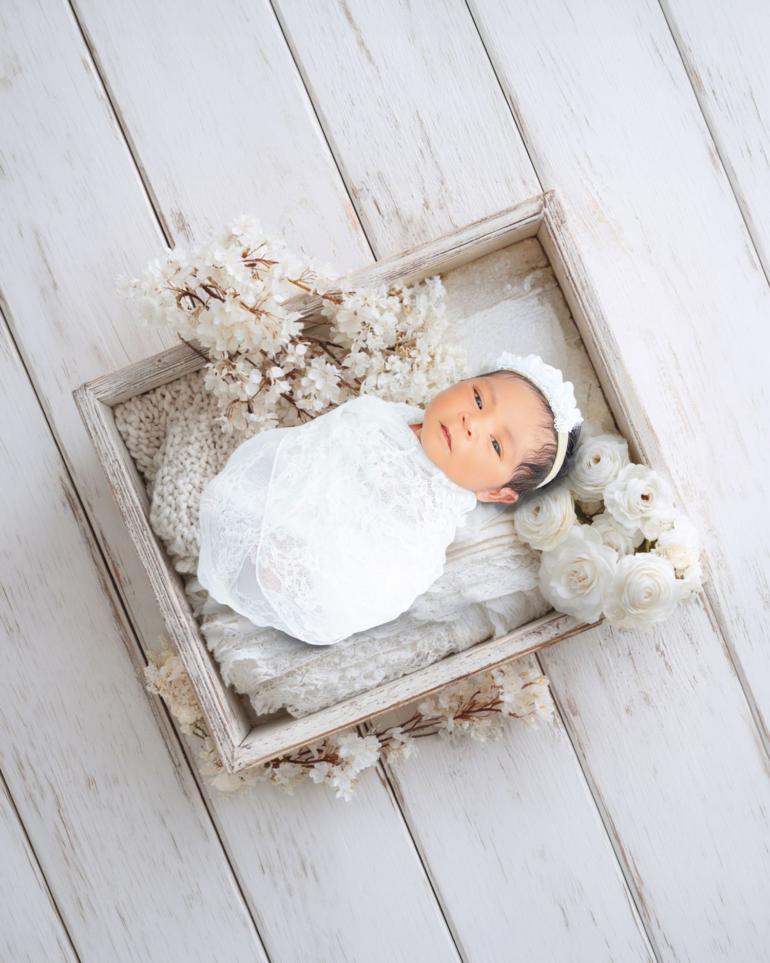 Newborn baby swaddled in white lace wrap resting in rustic wooden box with cream florals.