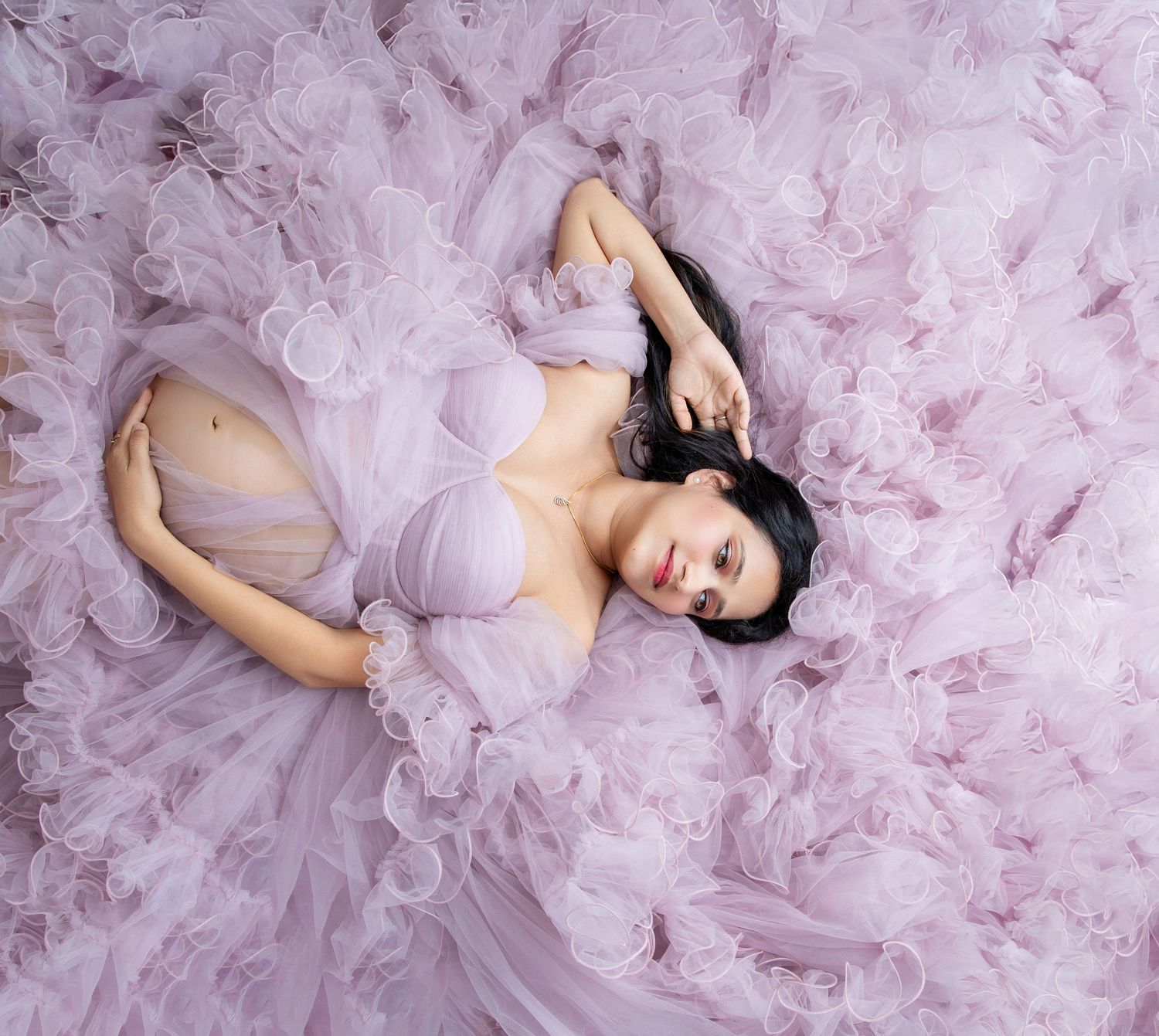 A pregnant woman in a lavender tulle gown lies gracefully surrounded by soft ruffled fabric in a dreamy maternity portrait.