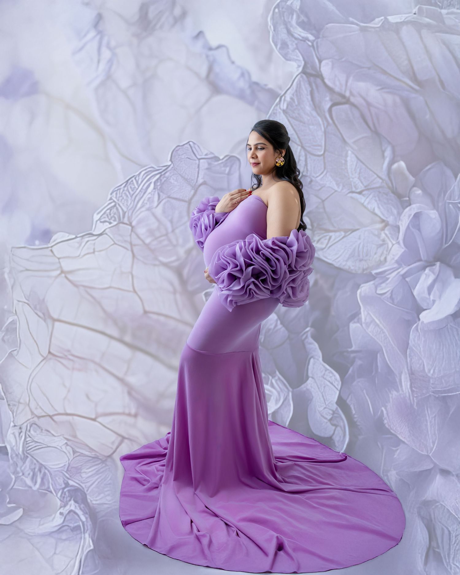 Pregnant woman in elegant lavender ruffle maternity gown poses gracefully against a floral white backdrop.