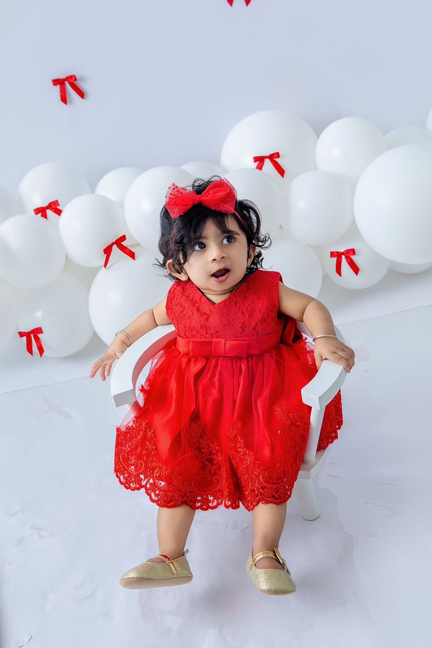Toddler in red dress and bow exploring white balloon decorated setting.