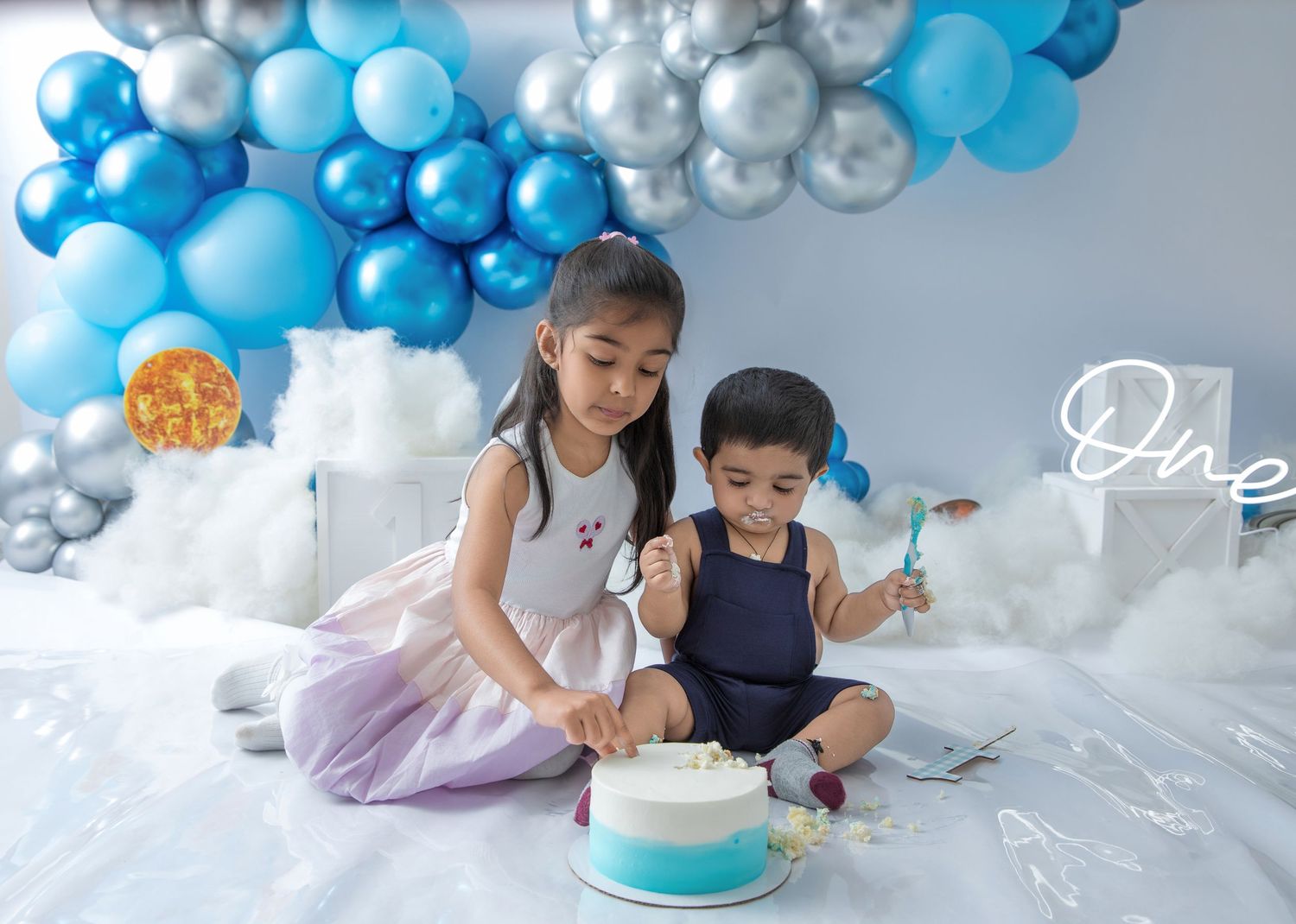 Baby in blue outfit plays with cake during cake smash photo session with balloon arch decorations.