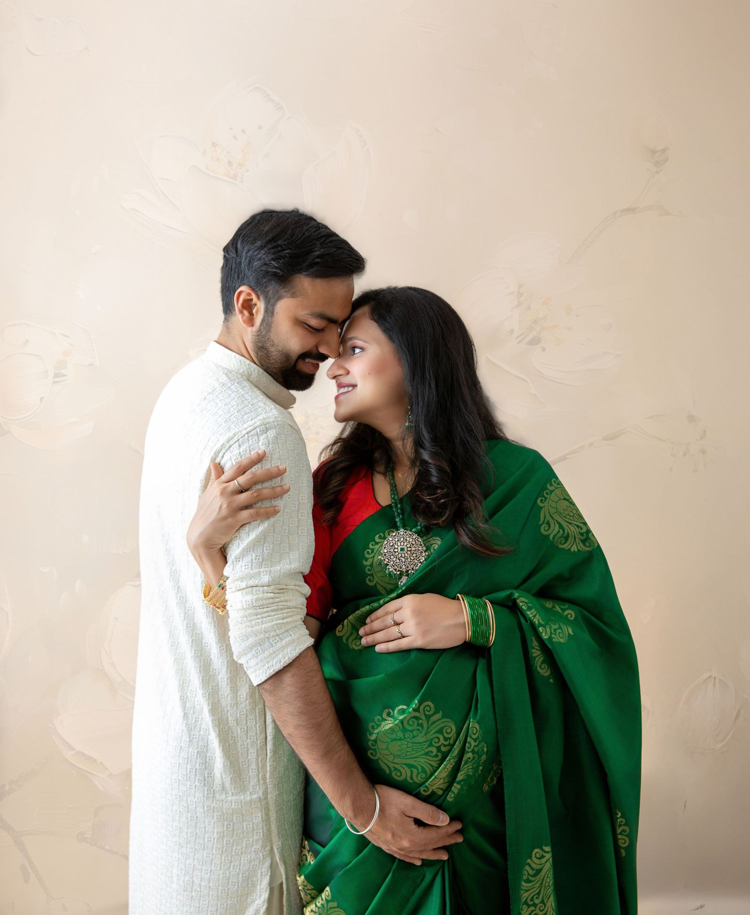 Pregnant Indian woman in green and red saree embraced by man in white kurta during maternity photoshoot.