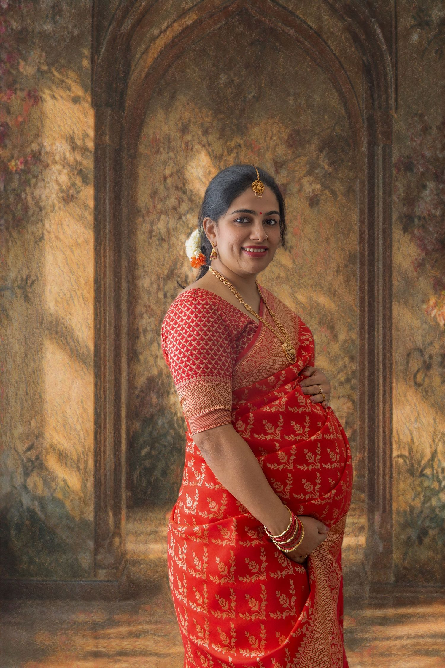 Pregnant Indian woman in red saree with gold jewelry smiling gracefully in front of ornate floral backdrop.