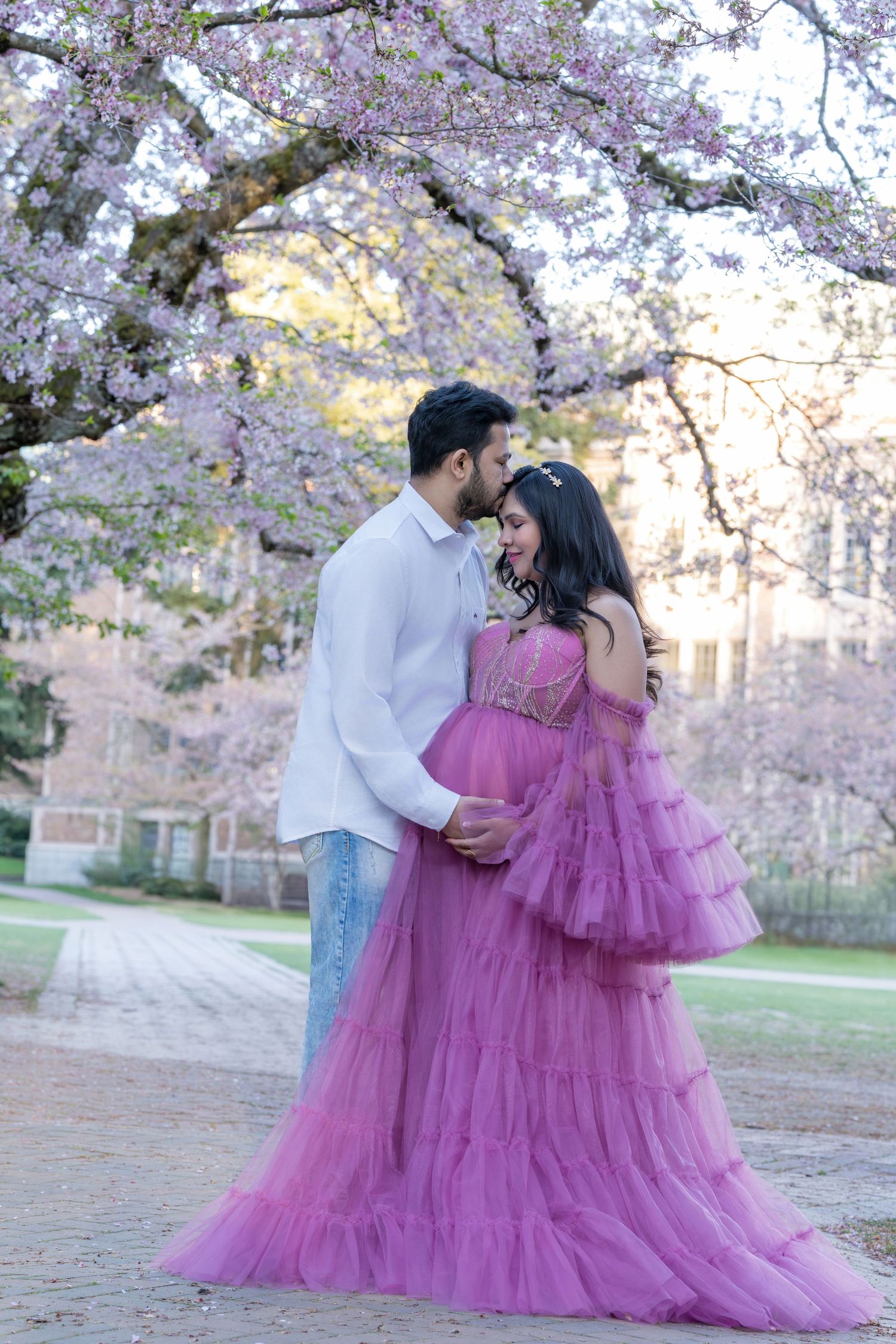 Couple sharing a tender moment under blooming cherry blossom trees, woman in elegant purple tulle maternity gown.