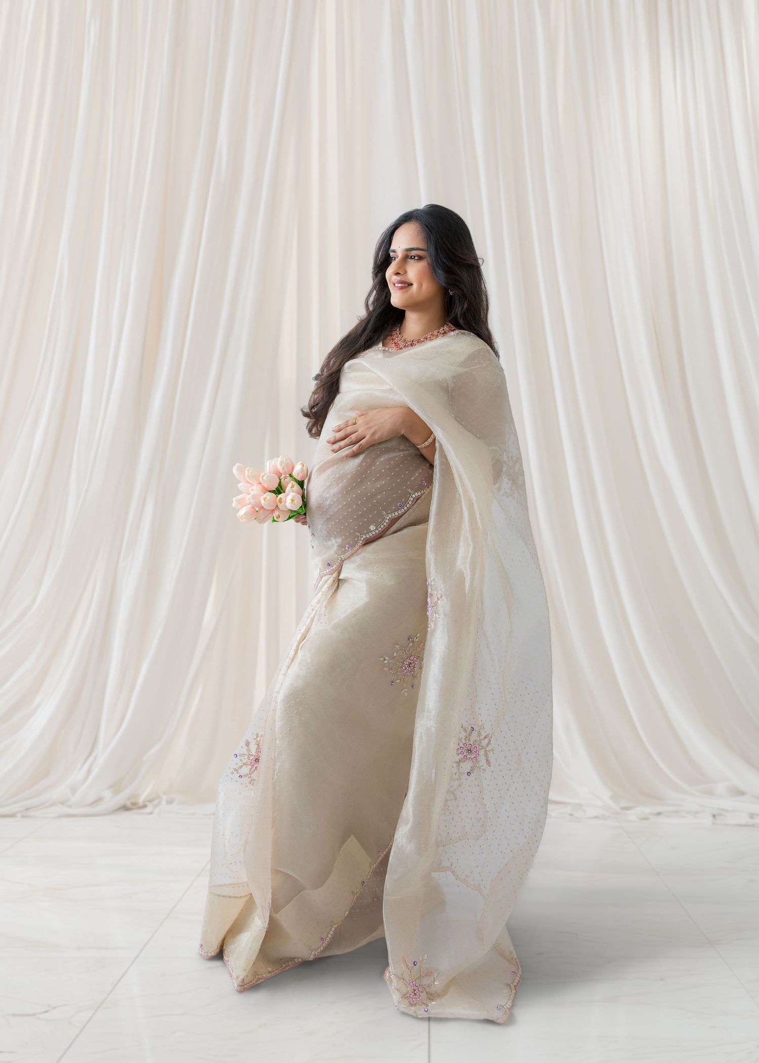 Pregnant woman in cream embroidered saree holds pink roses, smiling serenely before flowing white curtain backdrop.