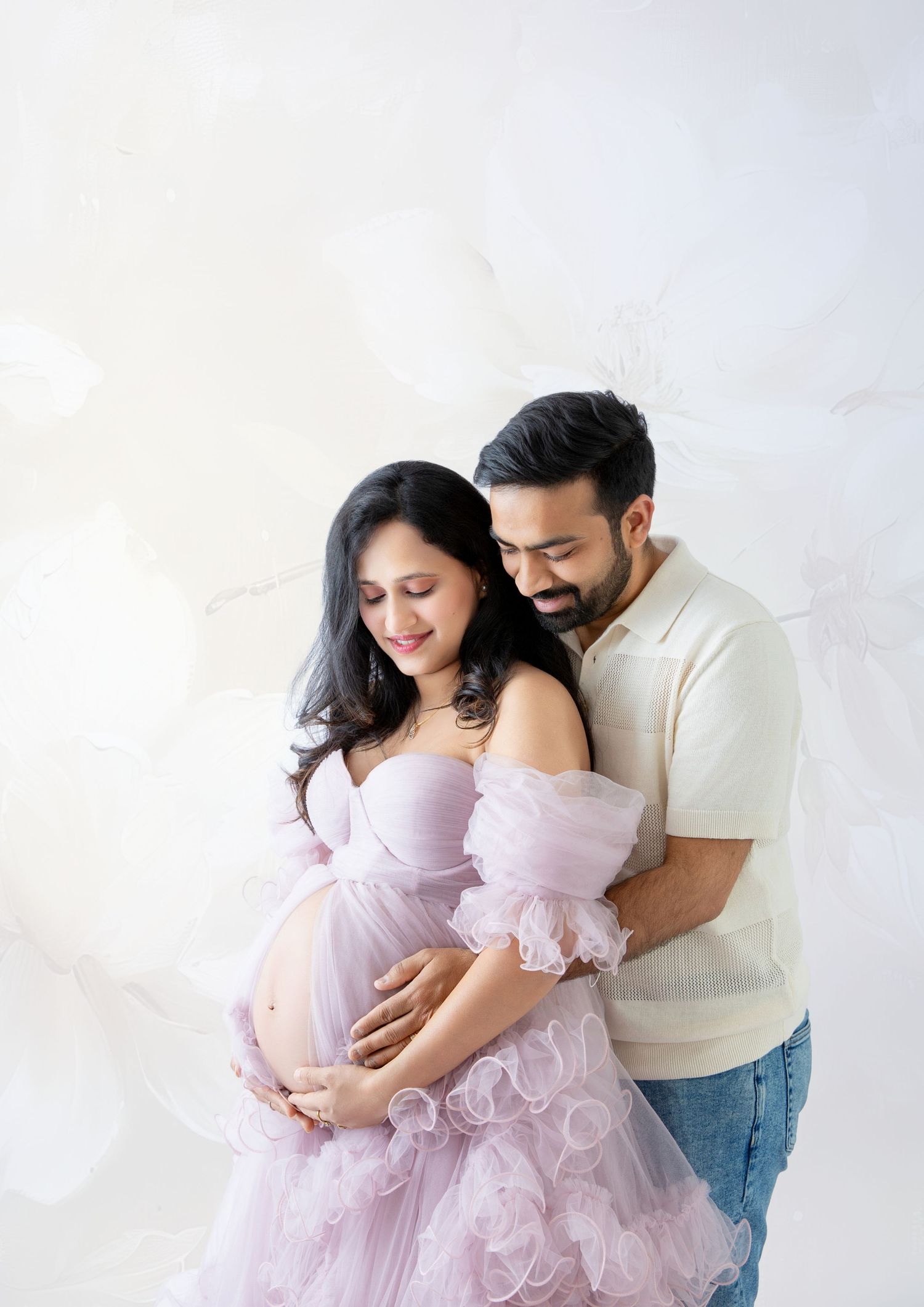 Couple in maternity photoshoot, woman in pink ruffle gown, man in cream shirt, both cradling baby bump.