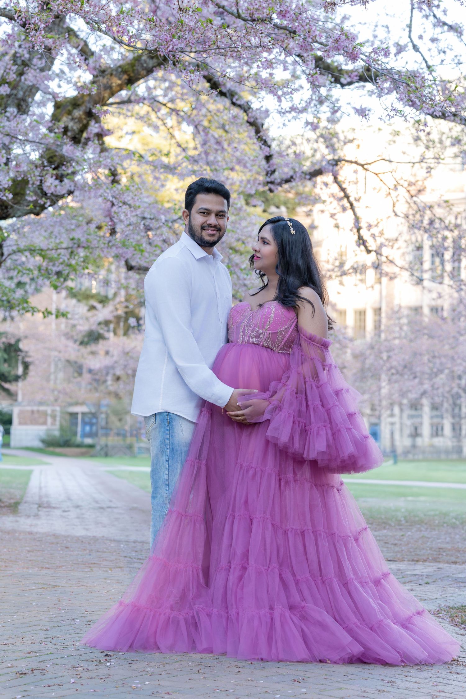 Pregnant couple posing under pink cherry blossom trees, woman in purple tulle gown, man in white shirt and jeans.