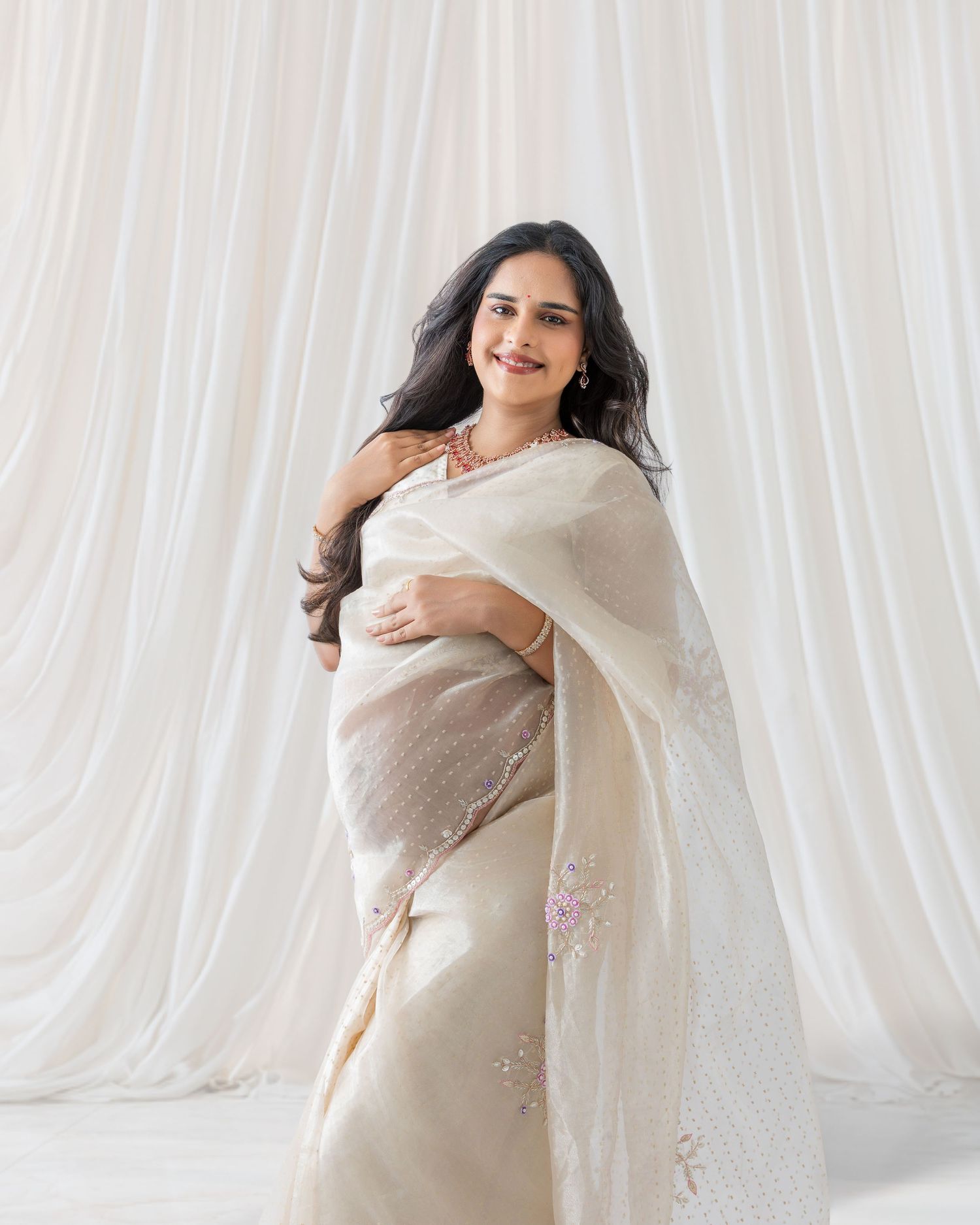Woman in elegant cream embroidered saree poses gracefully against white draped fabric backdrop.